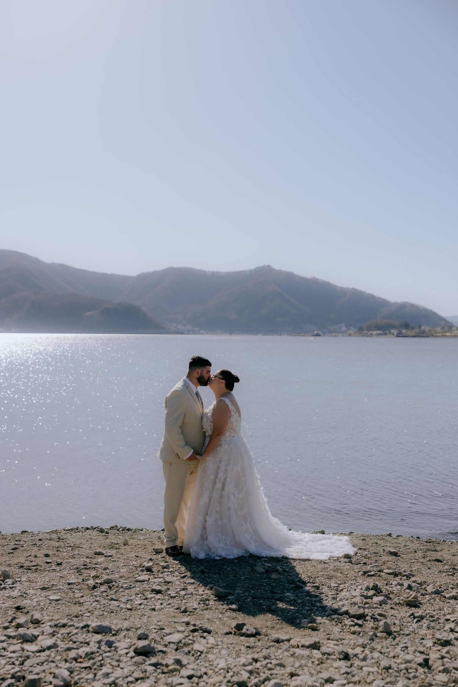 A romantic couple in wedding attire shares intimate moments by a scenic mountain lake shoreline at sunset.