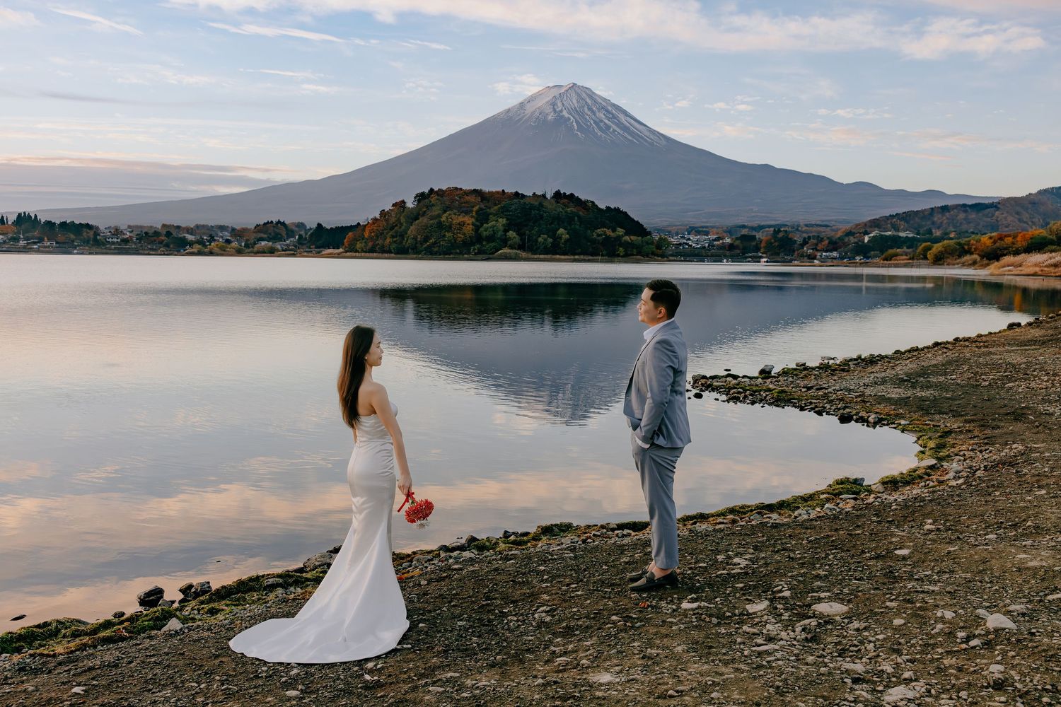 Two people stand at the edge of Lake Kawaguchiko with Mount Fuji rising majestically in the background at sunset.