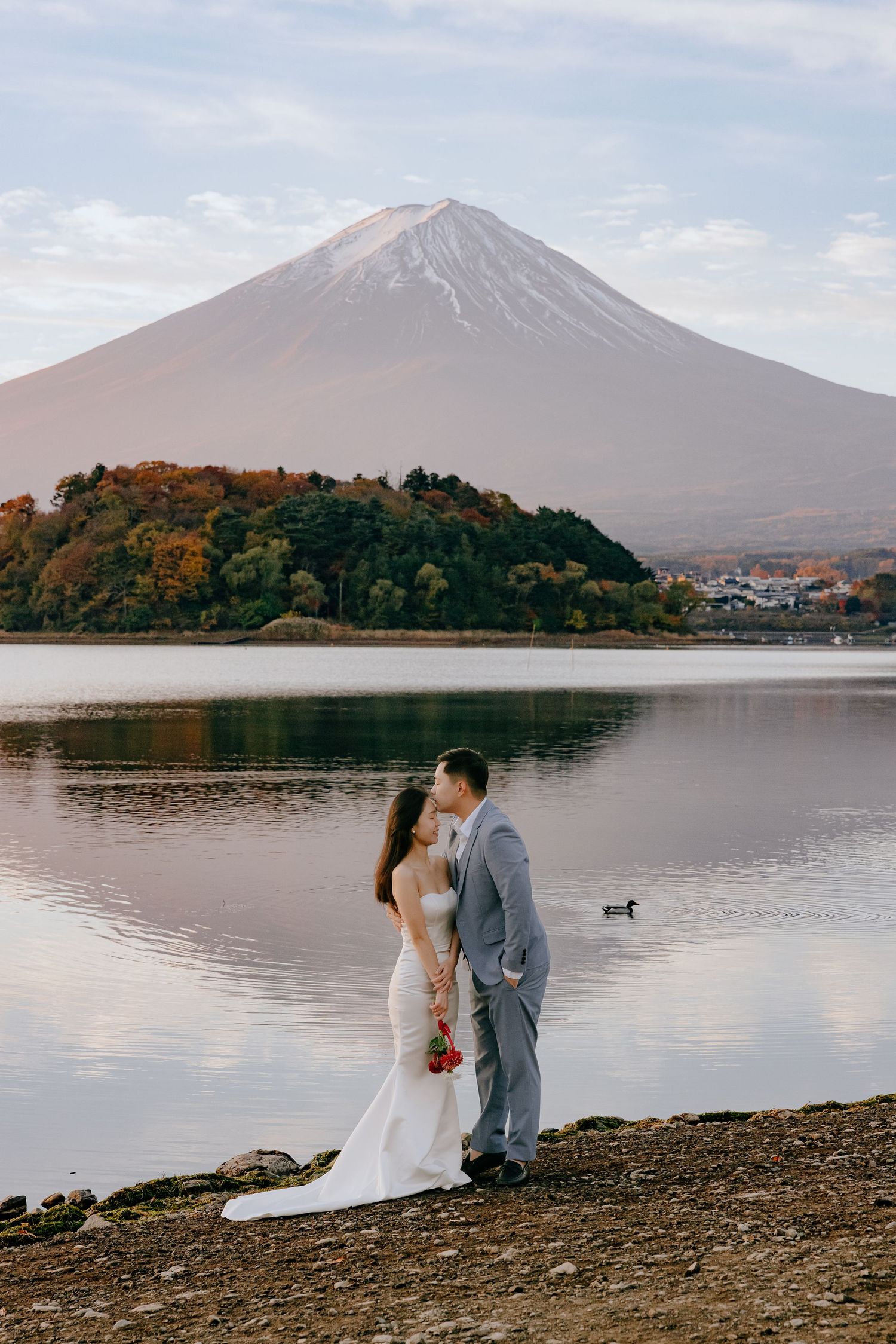 A couple in wedding attire sits by the serene Lake Kawaguchiko with Mount Fuji creating a majestic backdrop at sunset.