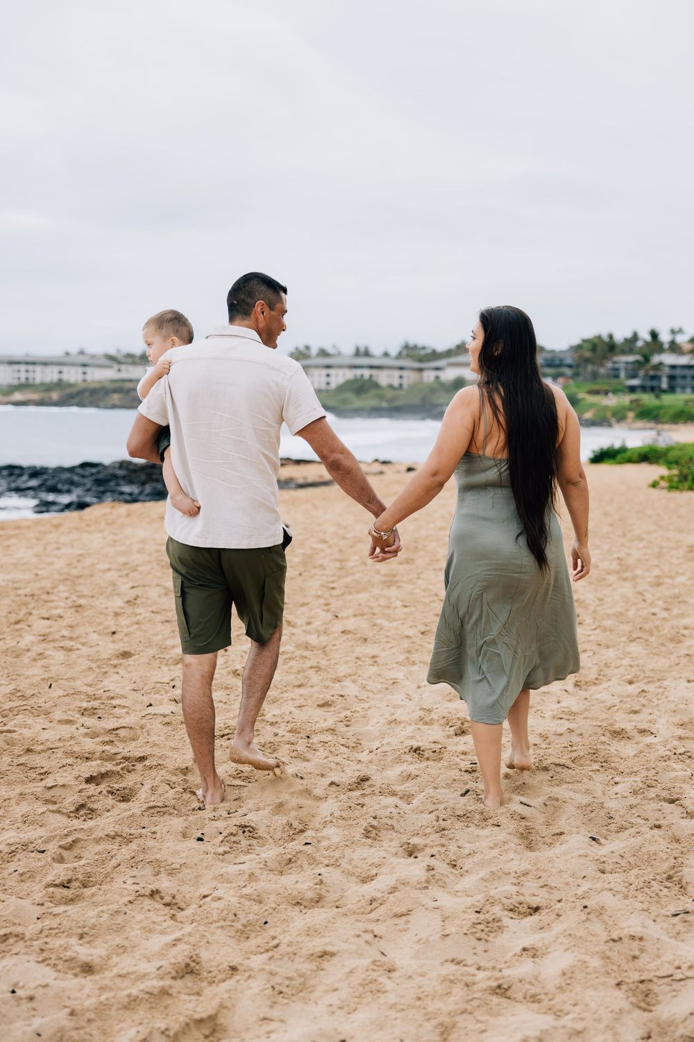 Family walking hand-in-hand on sandy beach, father carrying baby on shoulder.