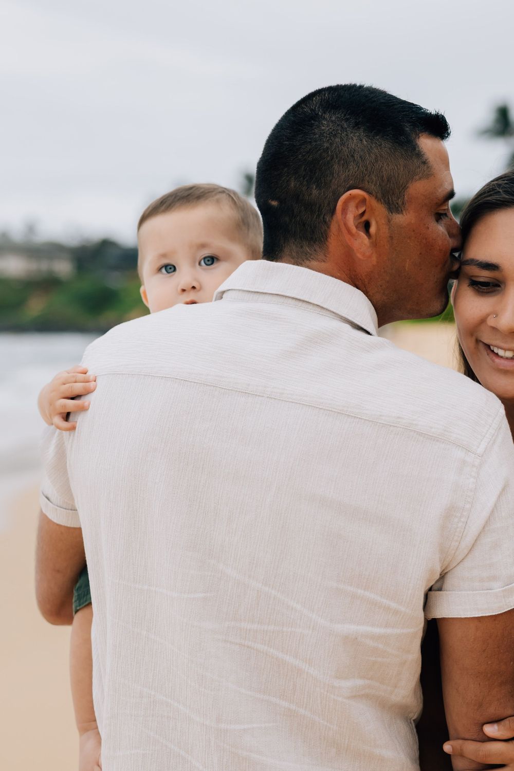 Baby with blue eyes peeks over father's shoulder as parents embrace on beach.
