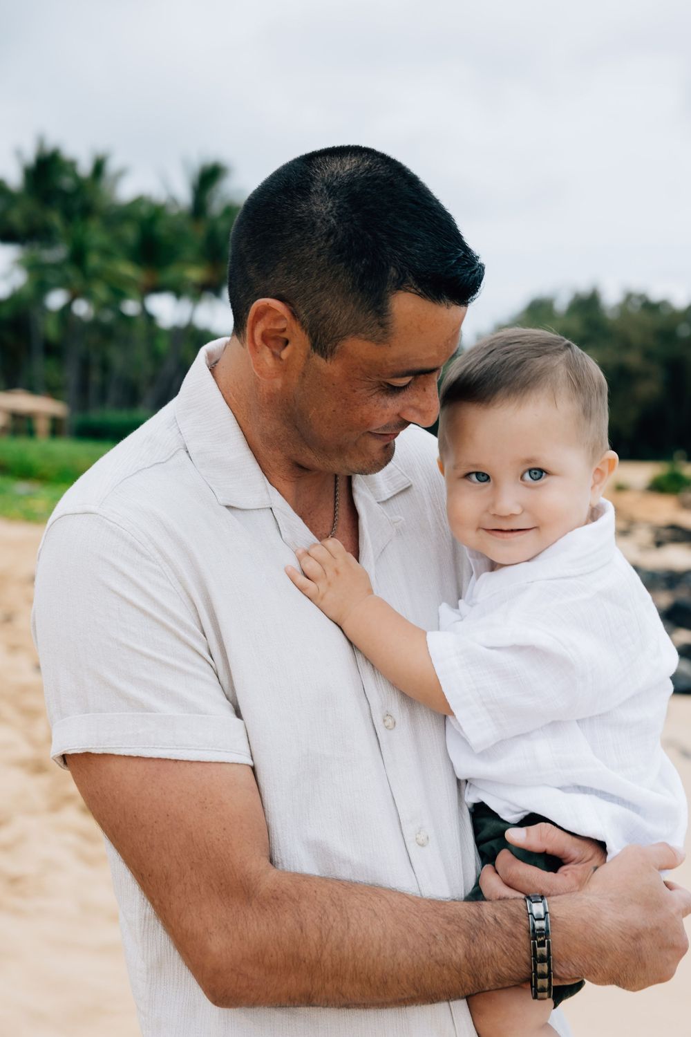 Father holding smiling baby boy on a sandy beach with palm trees.