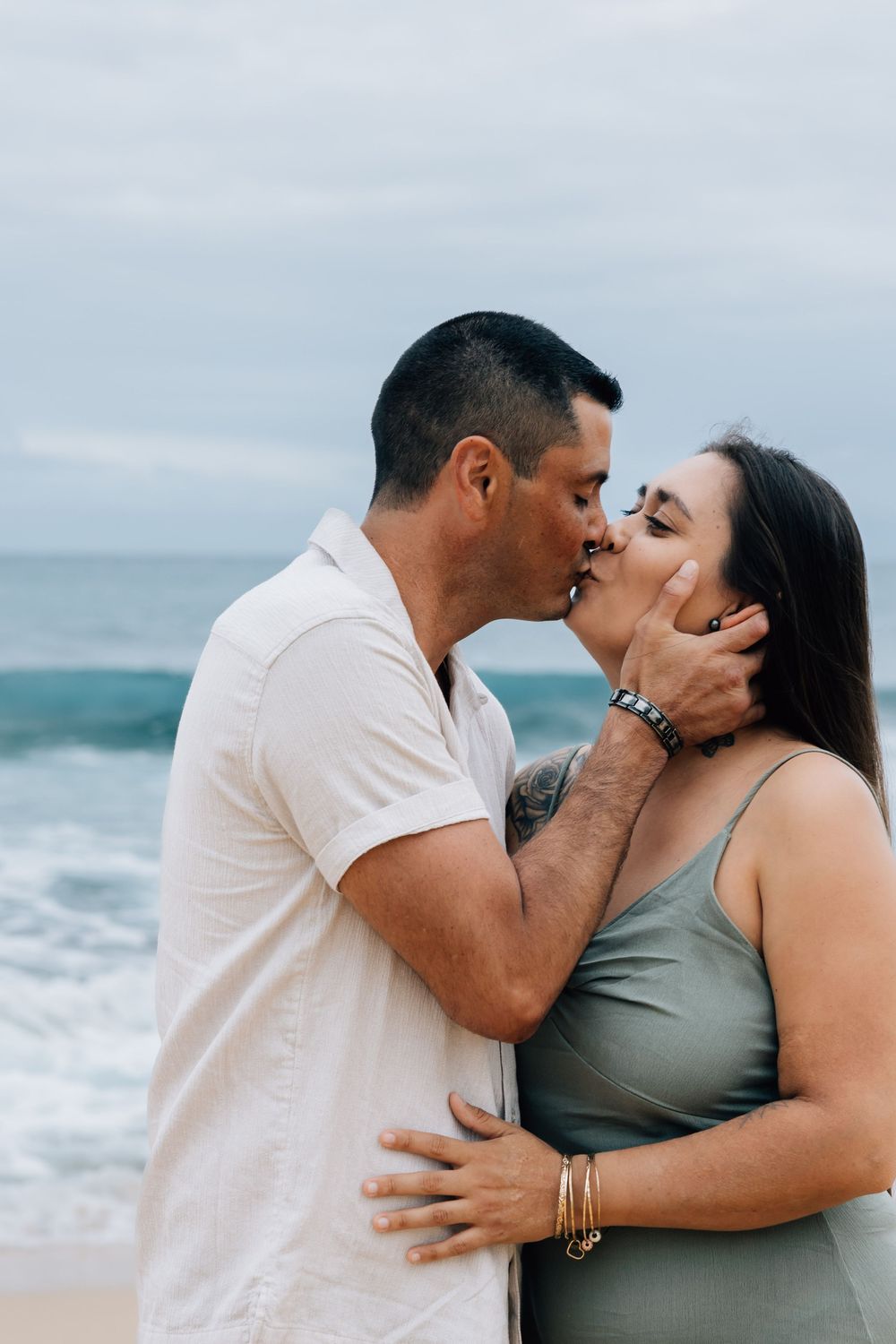 Couple sharing a romantic kiss on the beach with ocean waves behind them.