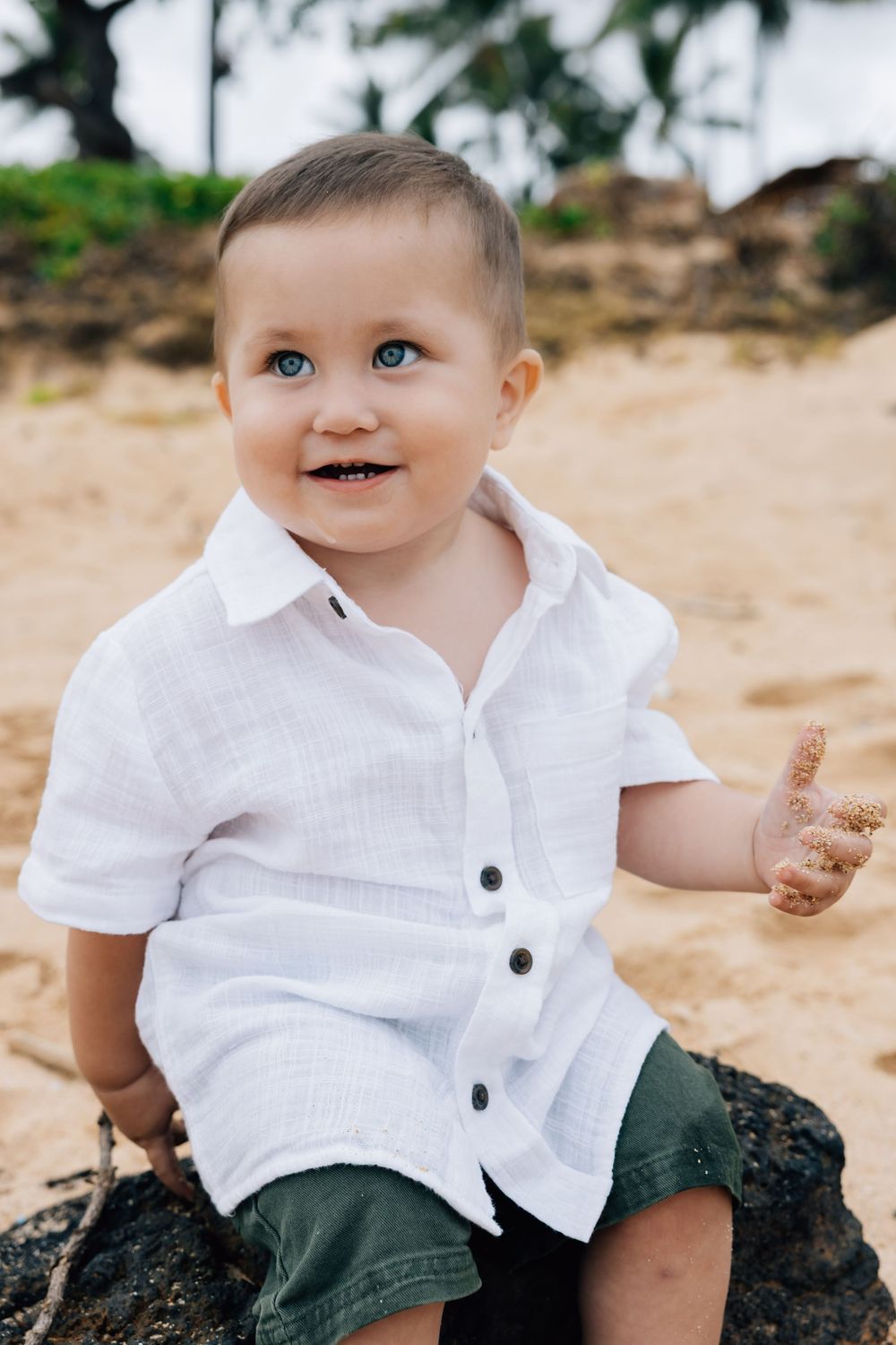 Adorable blue-eyed baby boy in white shirt sitting on rock at beach.