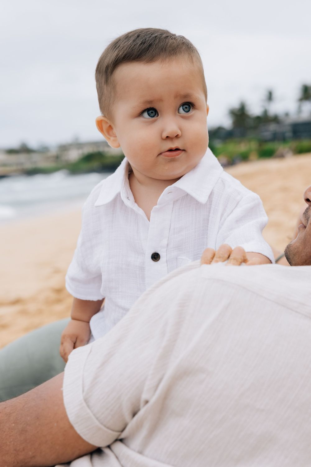 Adorable blue-eyed baby boy in white shirt sitting on beach.