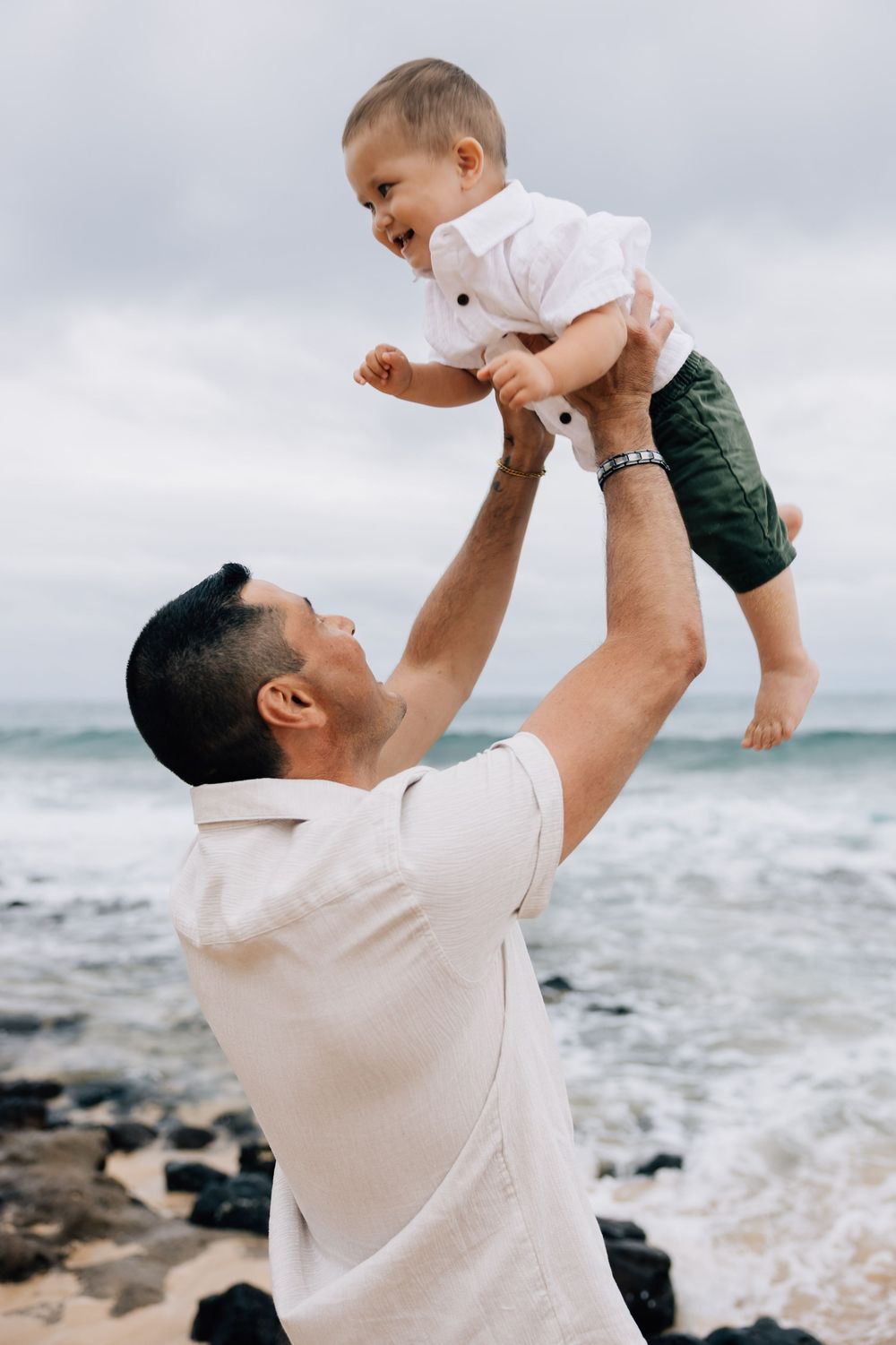 Father joyfully lifting laughing baby boy in the air at beach.