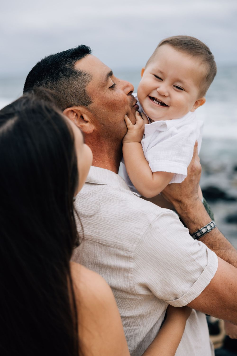 Parents kissing and cuddling their giggling baby boy at the beach.