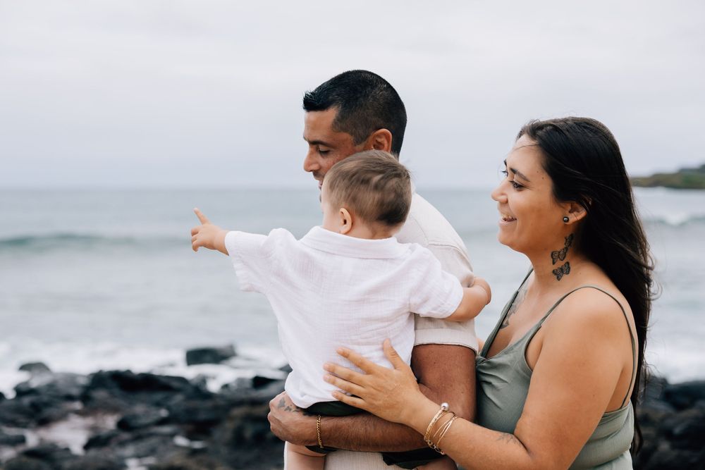 Young family with baby pointing at ocean view on rocky beach.
