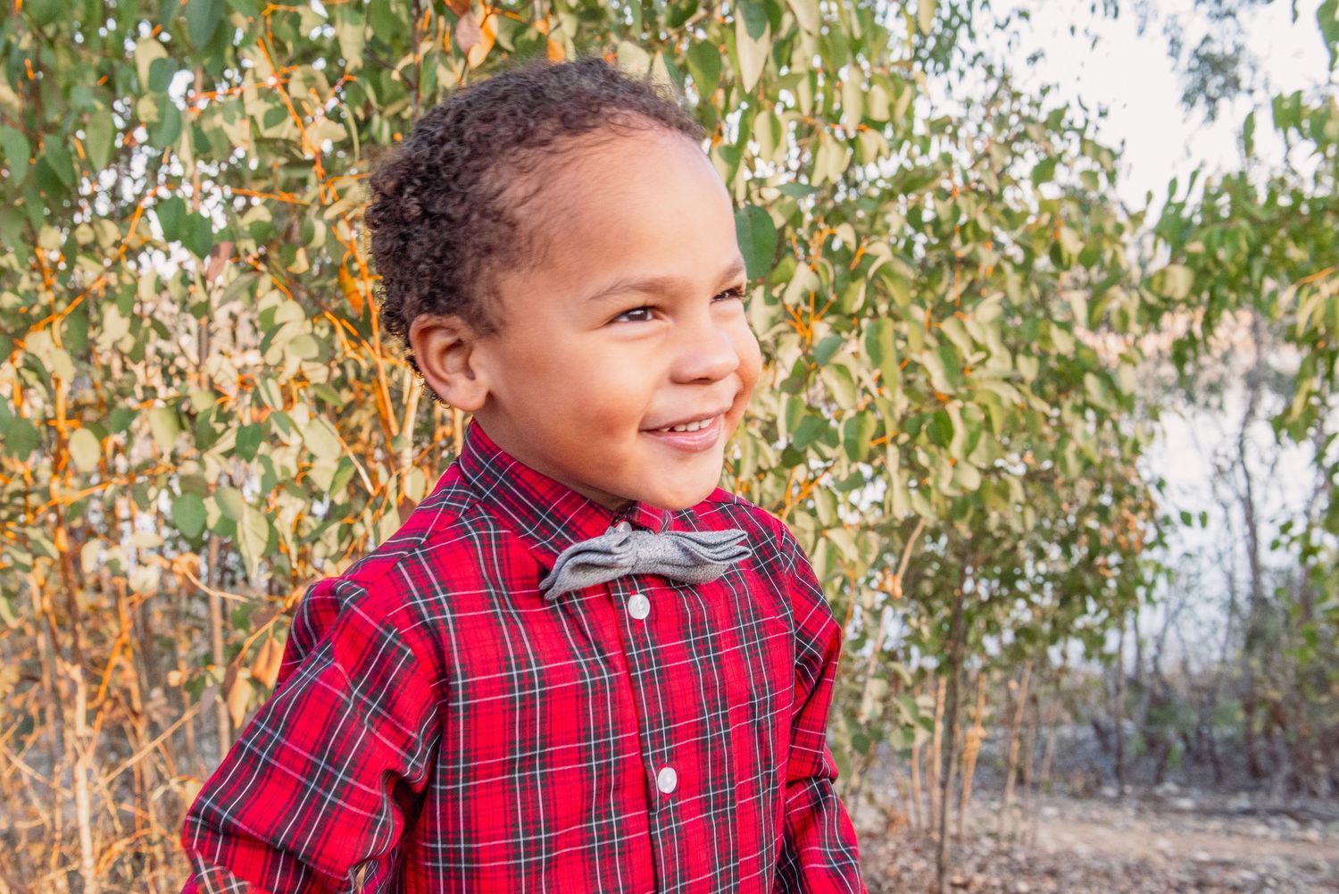 A person in a red plaid shirt smiles joyfully while posing outdoors against a natural autumn background.
