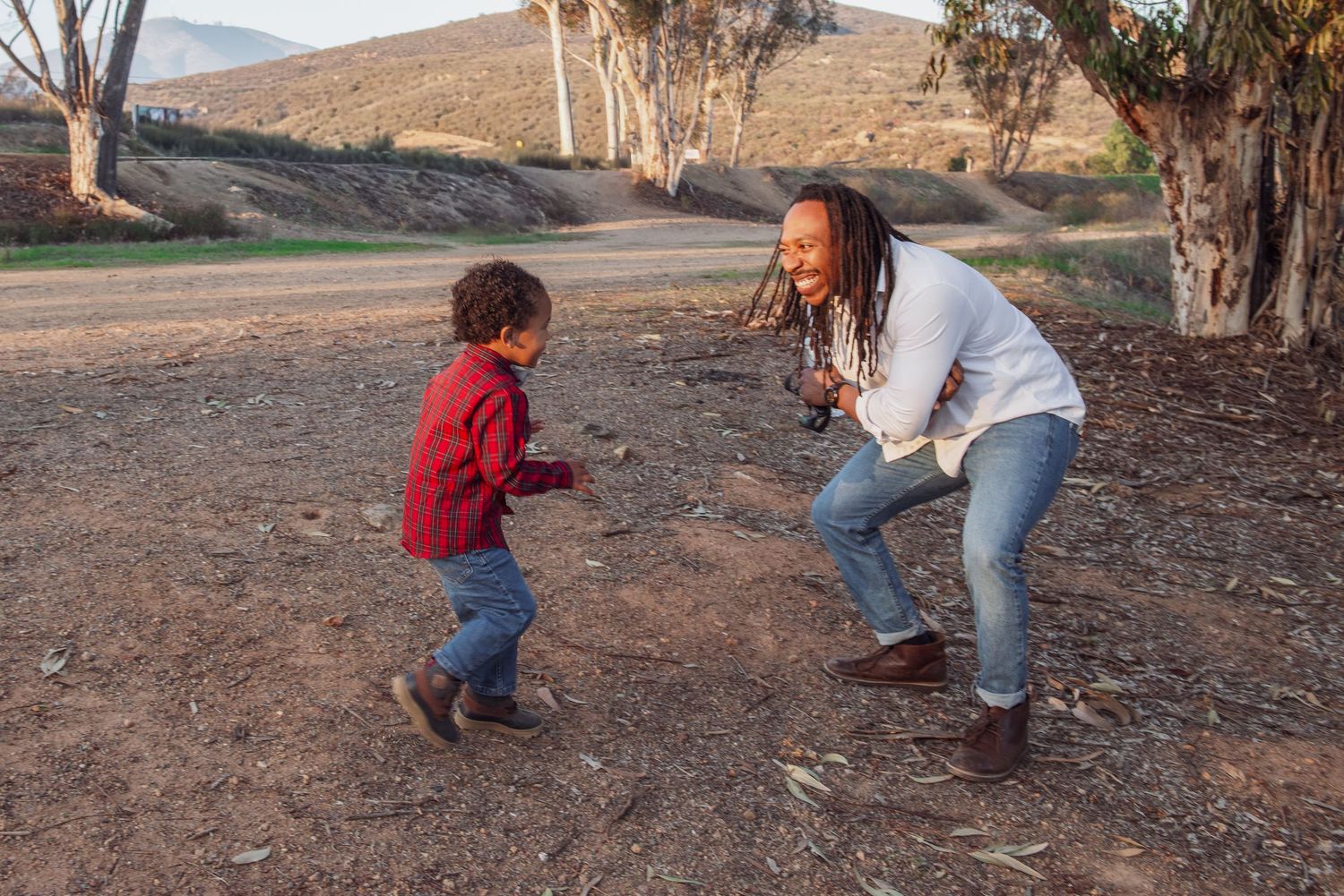 Two people playing and dancing together on a dirt path with mountains and trees in the background at sunset.