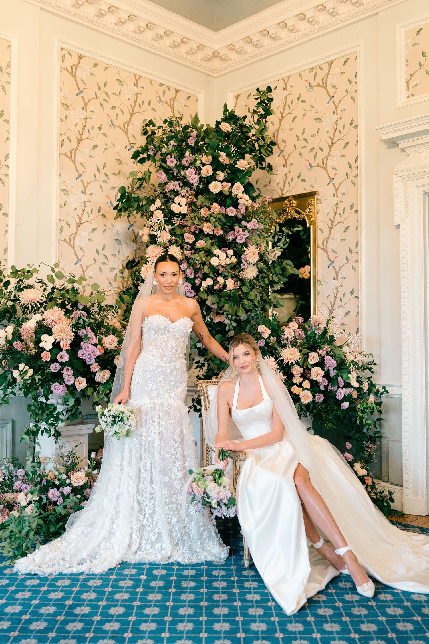 Two wedding dresses displayed in an elegant room with ornate wallpaper and a stunning pink rose floral arch installation.