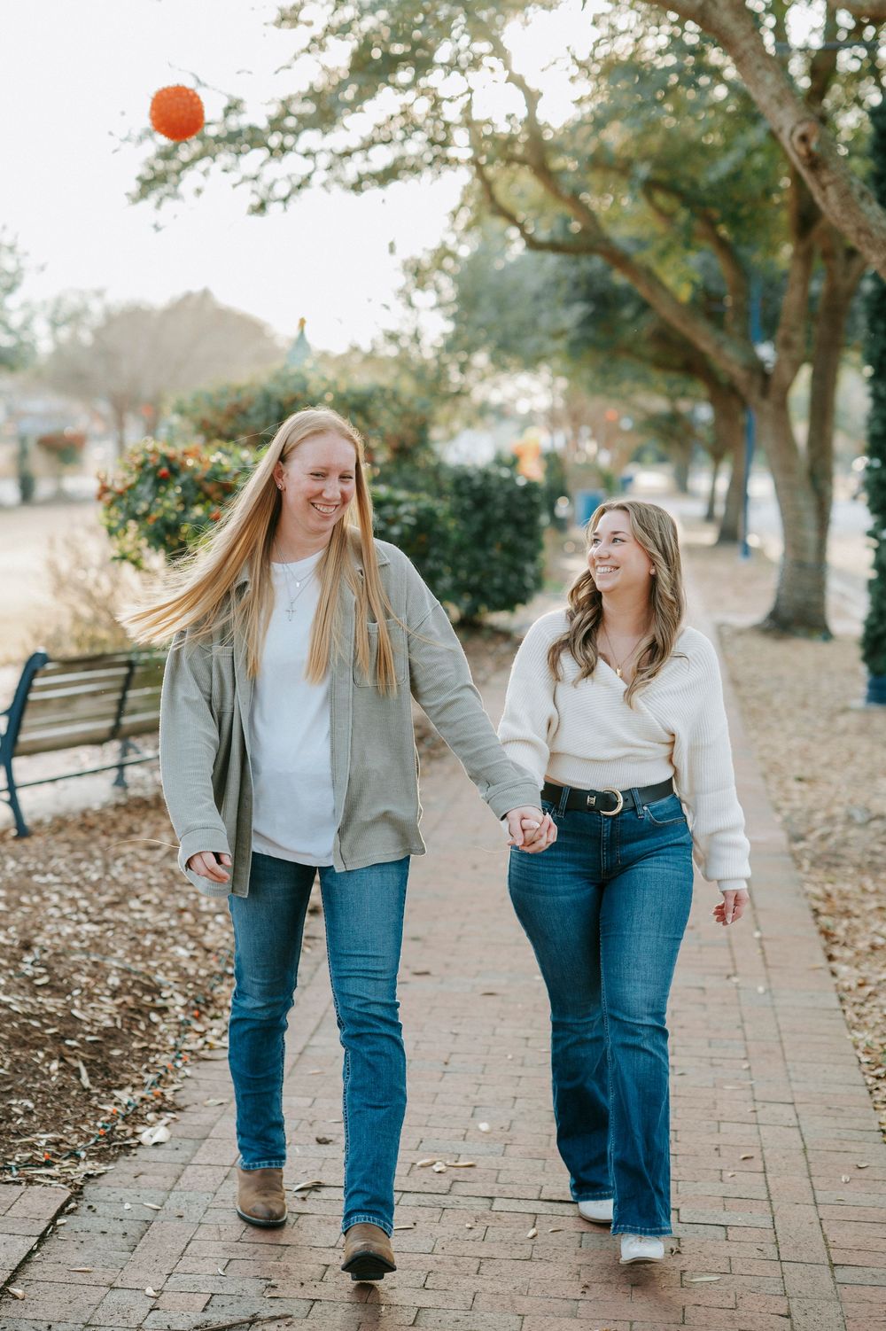 Two people in matching jeans and sweaters playfully walk together on a brick path surrounded by autumn leaves.