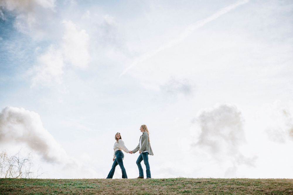Two people walk hand in hand along a grassy hilltop silhouetted against a cloudy sky at sunset.