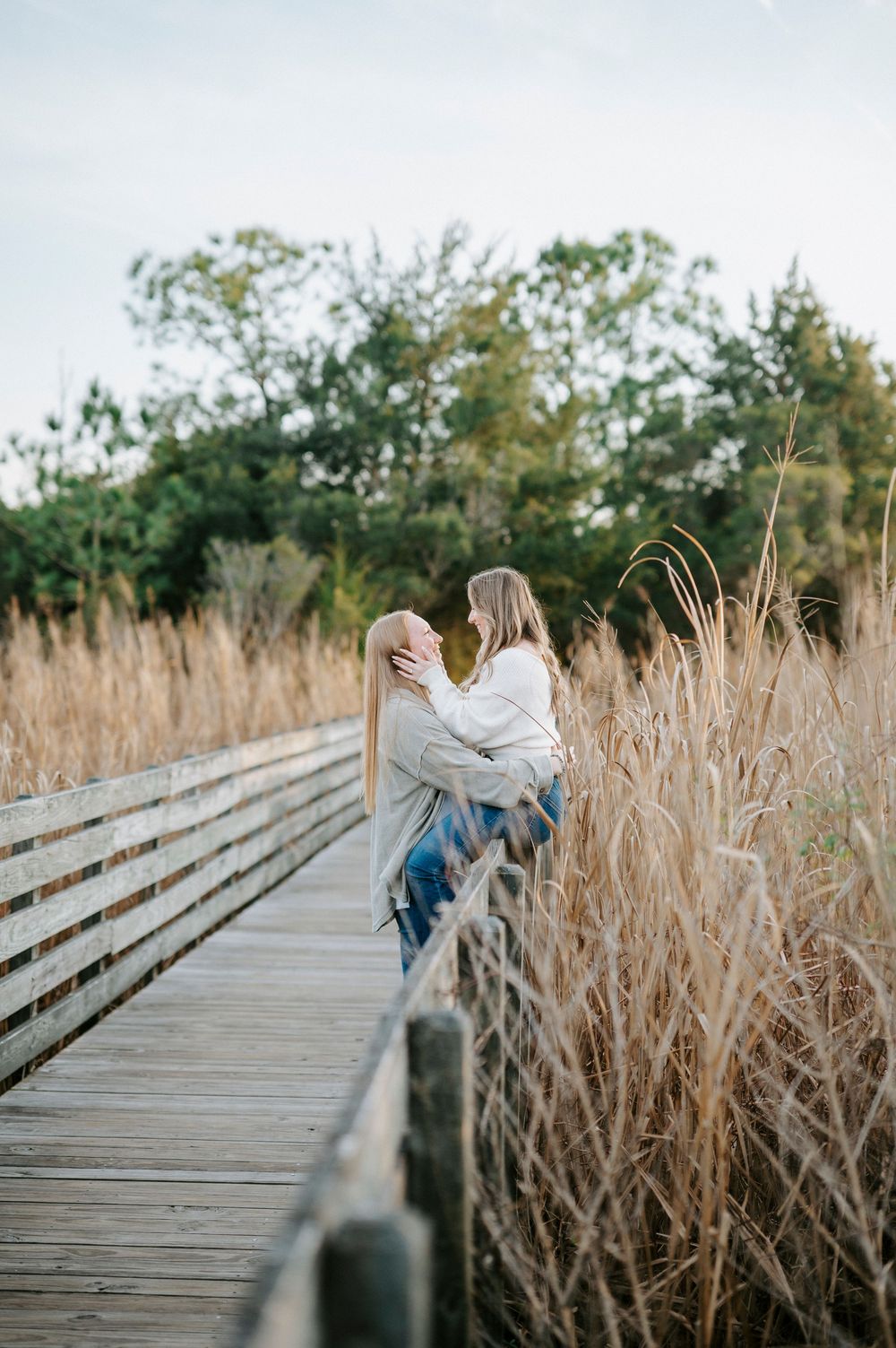 A couple embraces on a wooden boardwalk surrounded by tall dried grasses during golden hour.