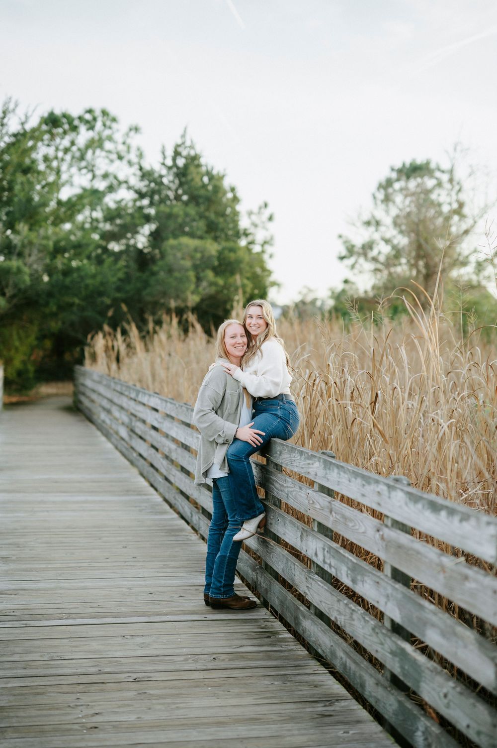 Two people sit close together on a wooden boardwalk railing with marsh grass in the background.