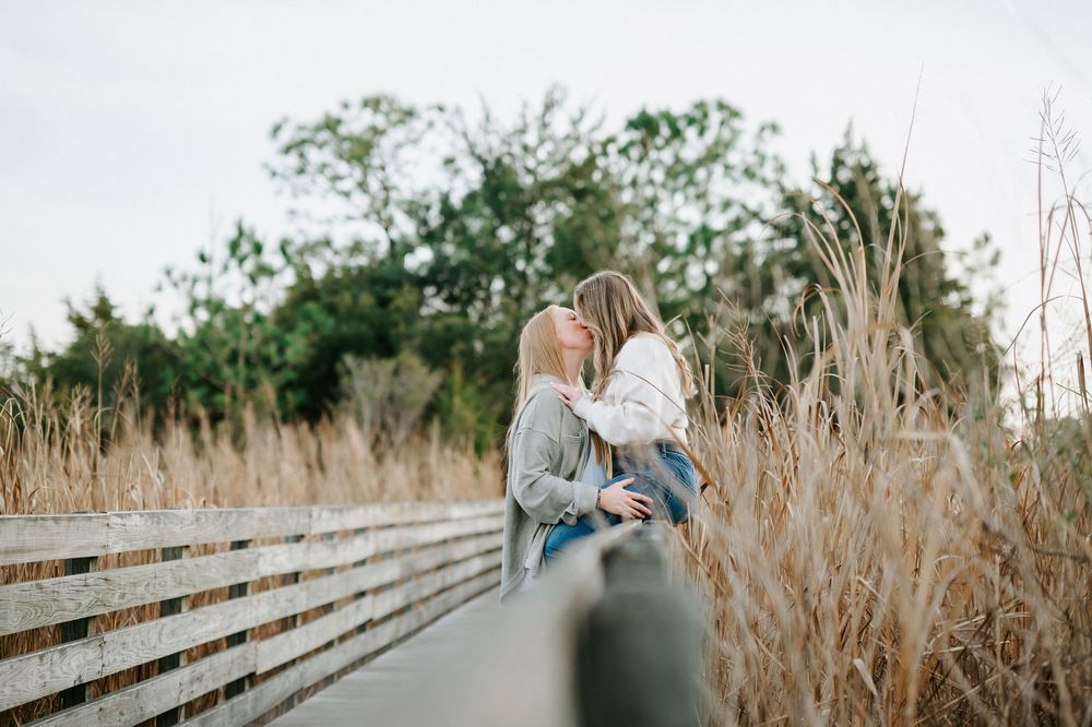 Couple relaxing on a rustic wooden fence surrounded by tall dried grass in a natural outdoor setting.