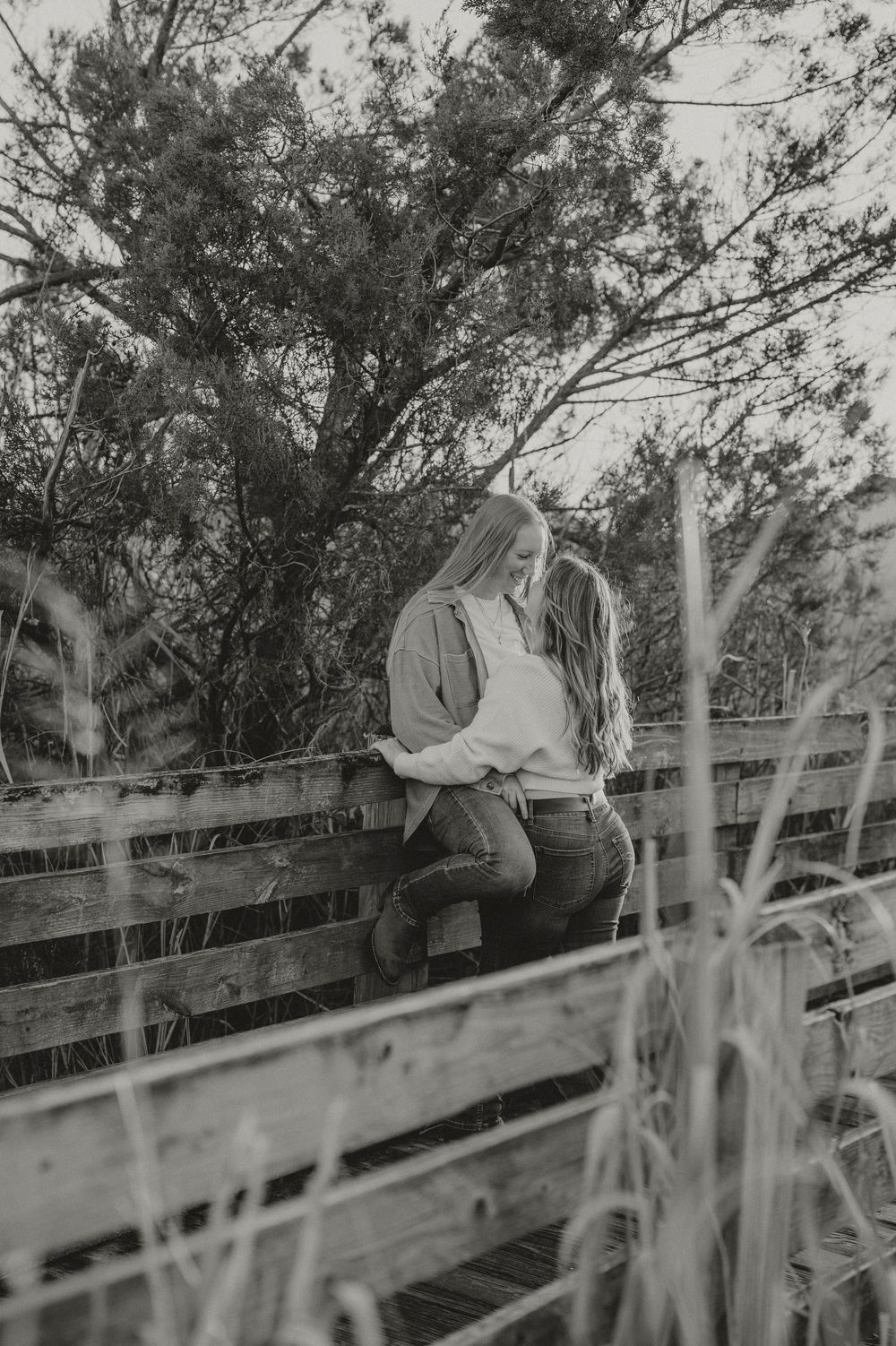 A vintage black and white photograph shows someone sitting casually on a wooden fence in a rural setting.