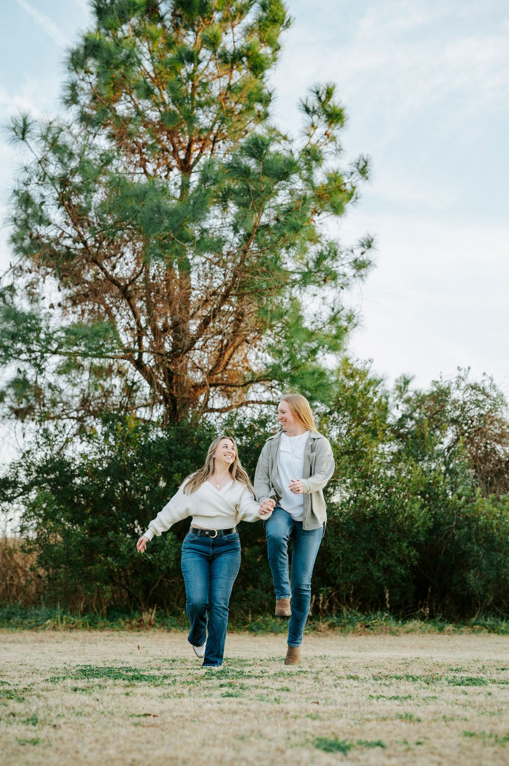 Two people in matching outfits laugh and play together in an open field with pine trees.