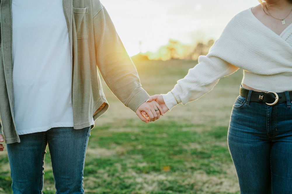 Couple holding hands during sunset in a grassy field wearing casual denim and sweaters.