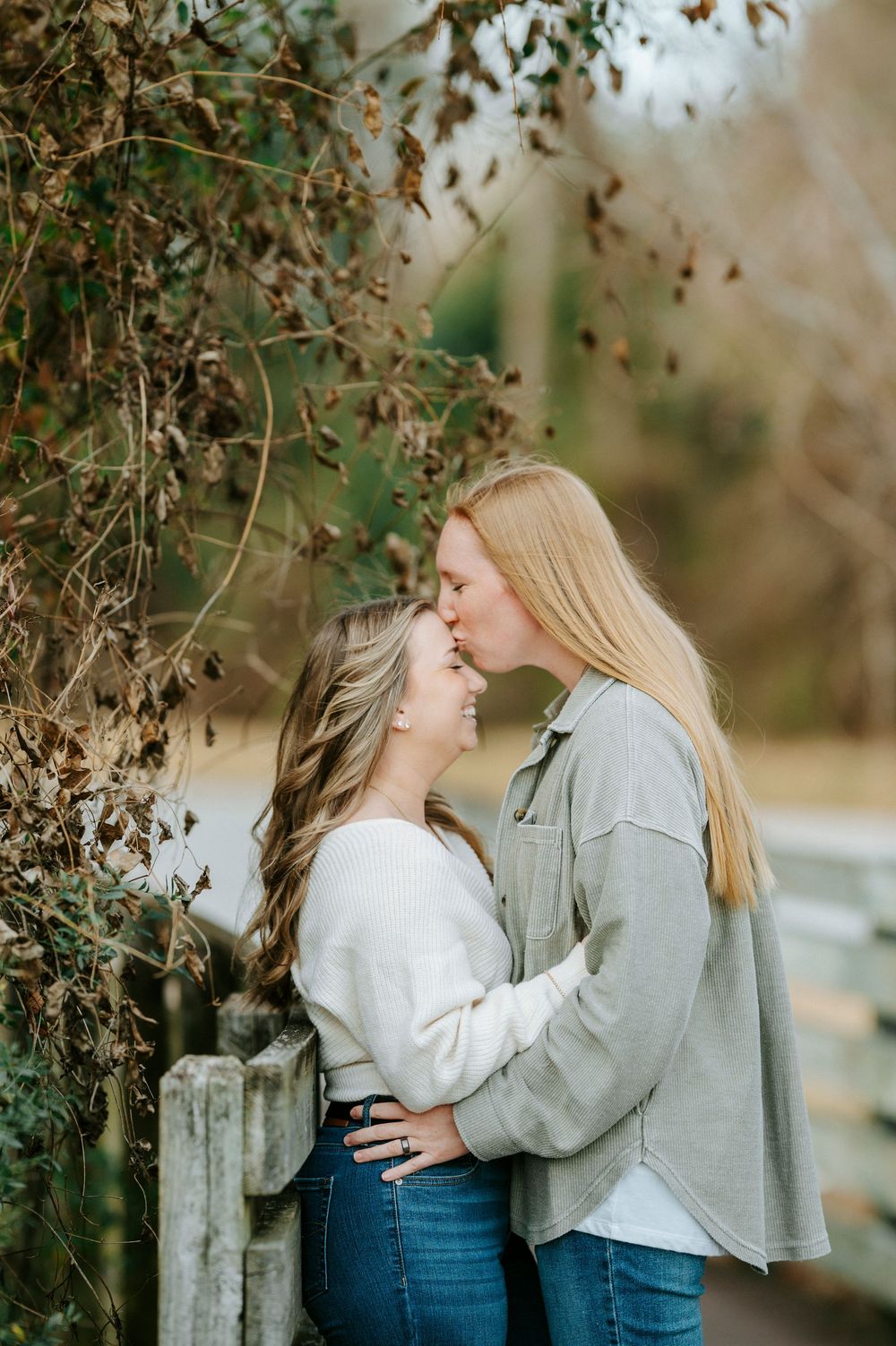 Two people in coordinated outfits share an intimate moment while standing near trees in natural lighting.