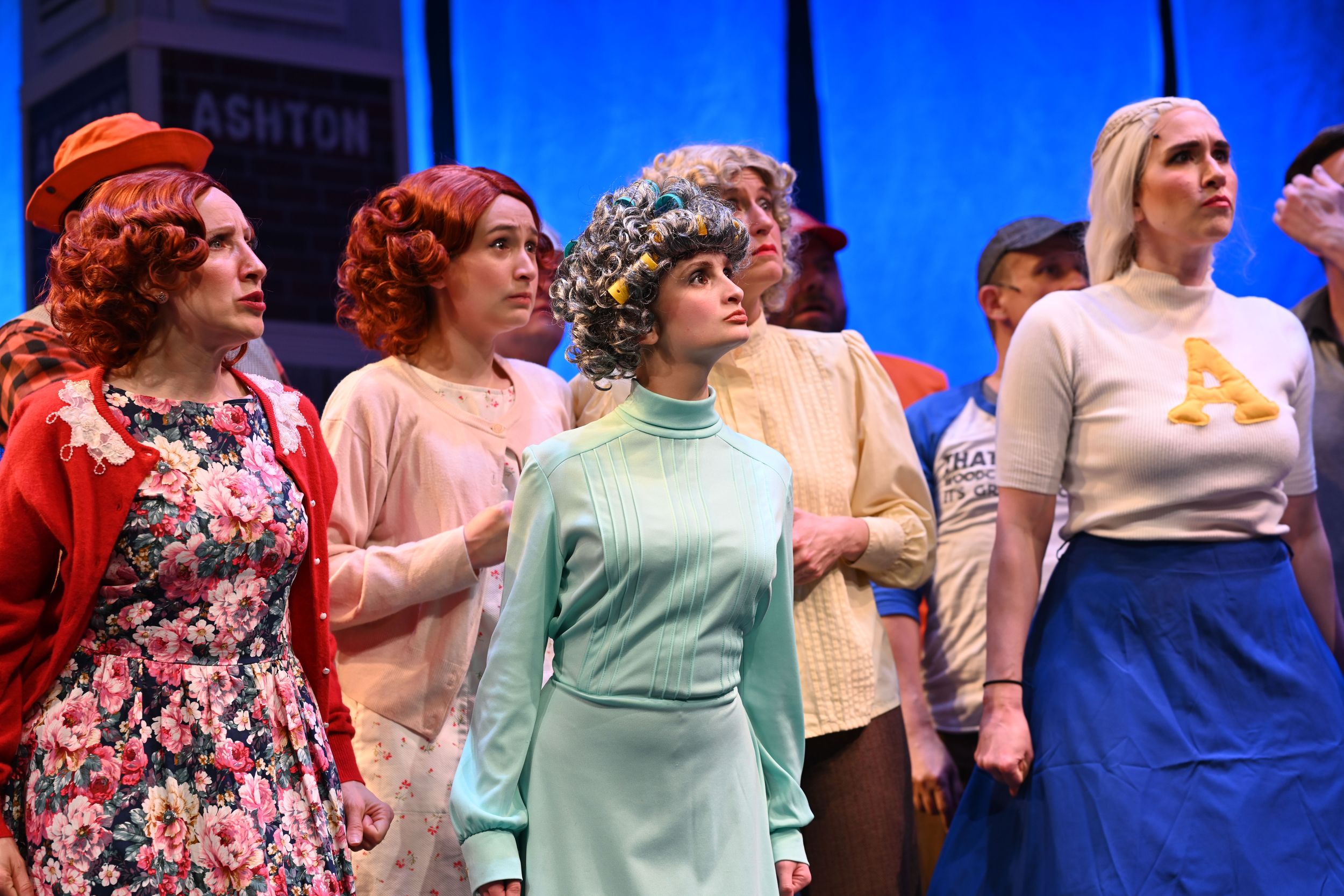 Theater ensemble cast performing in colorful 1950s style costumes against a bright blue backdrop.