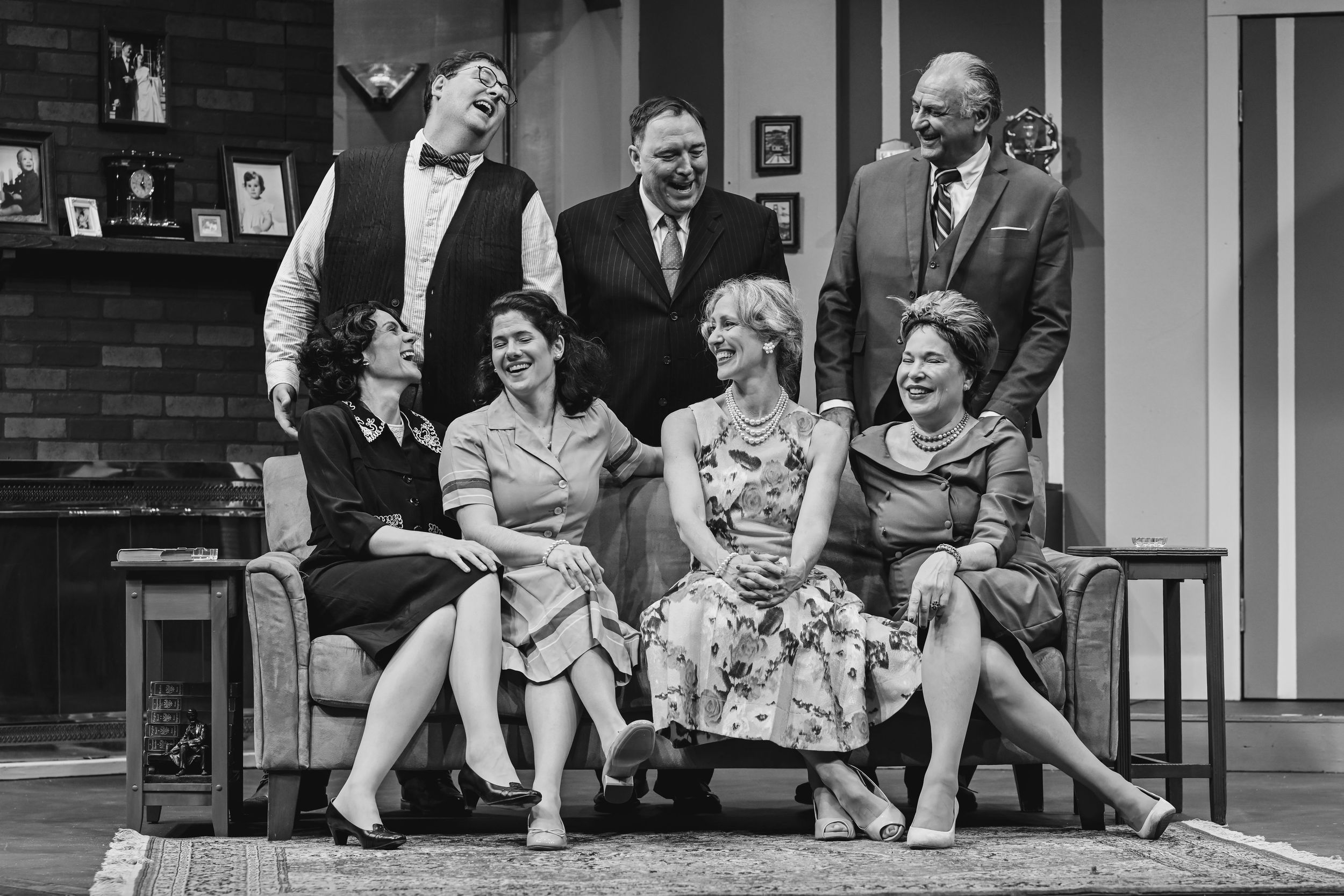 Black and white photograph of a theatrical living room scene with performers seated on furniture.