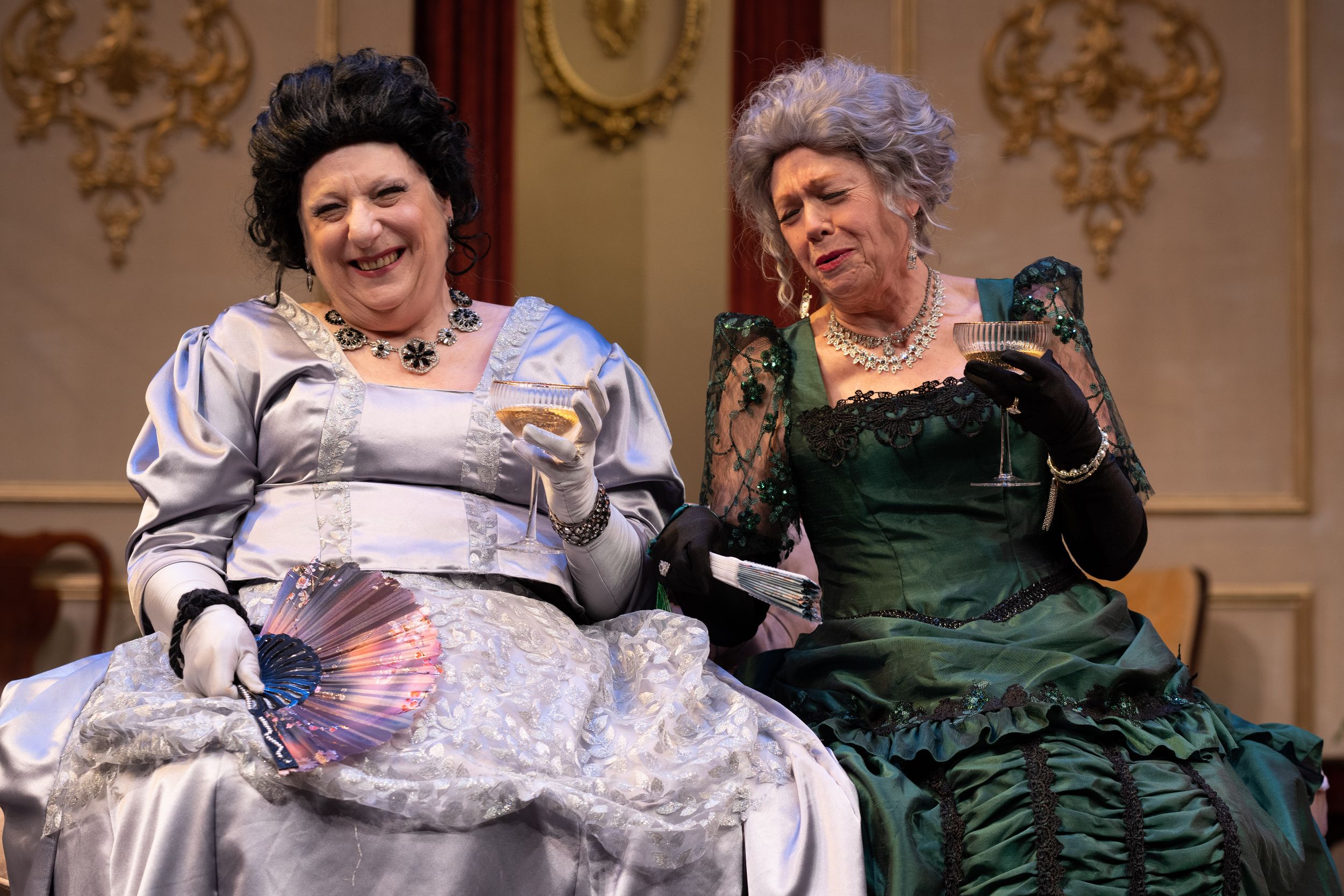 Theater scene with performers in elaborate Victorian-era gowns on ornate stage set.
