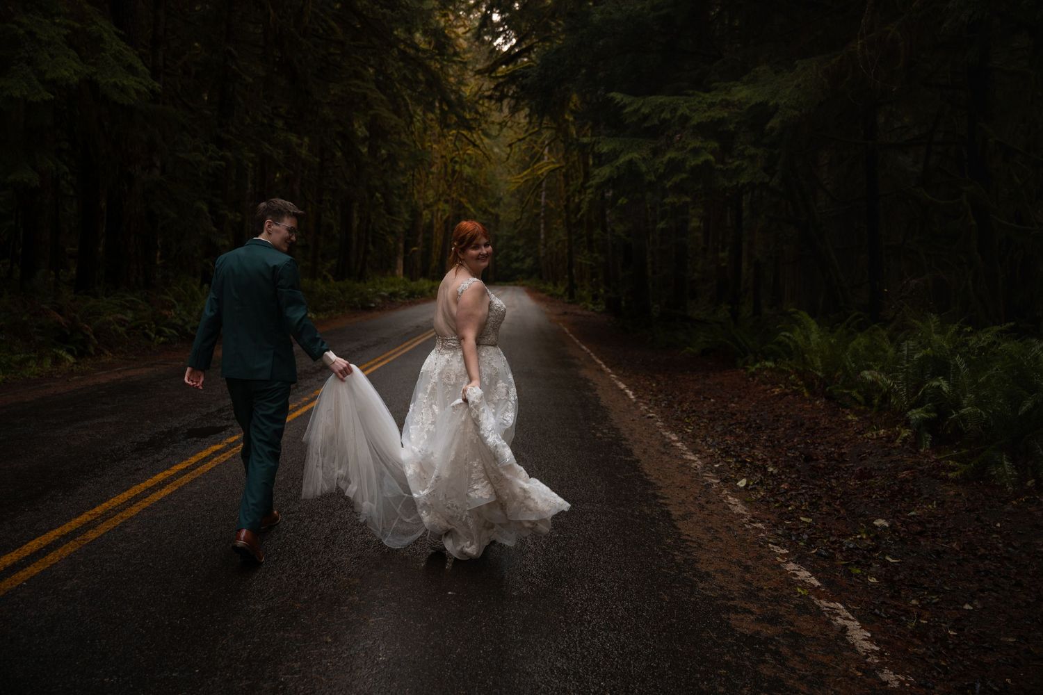 A bride walks down an empty road while her wife carries the train of her dress surrounded by ferns and mossy trees in the Pacific Northwest