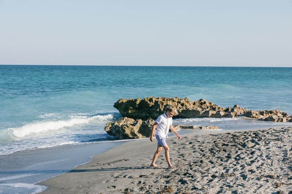 A child walks along a sandy beach near rocky shores with turquoise ocean waves under a clear blue sky.