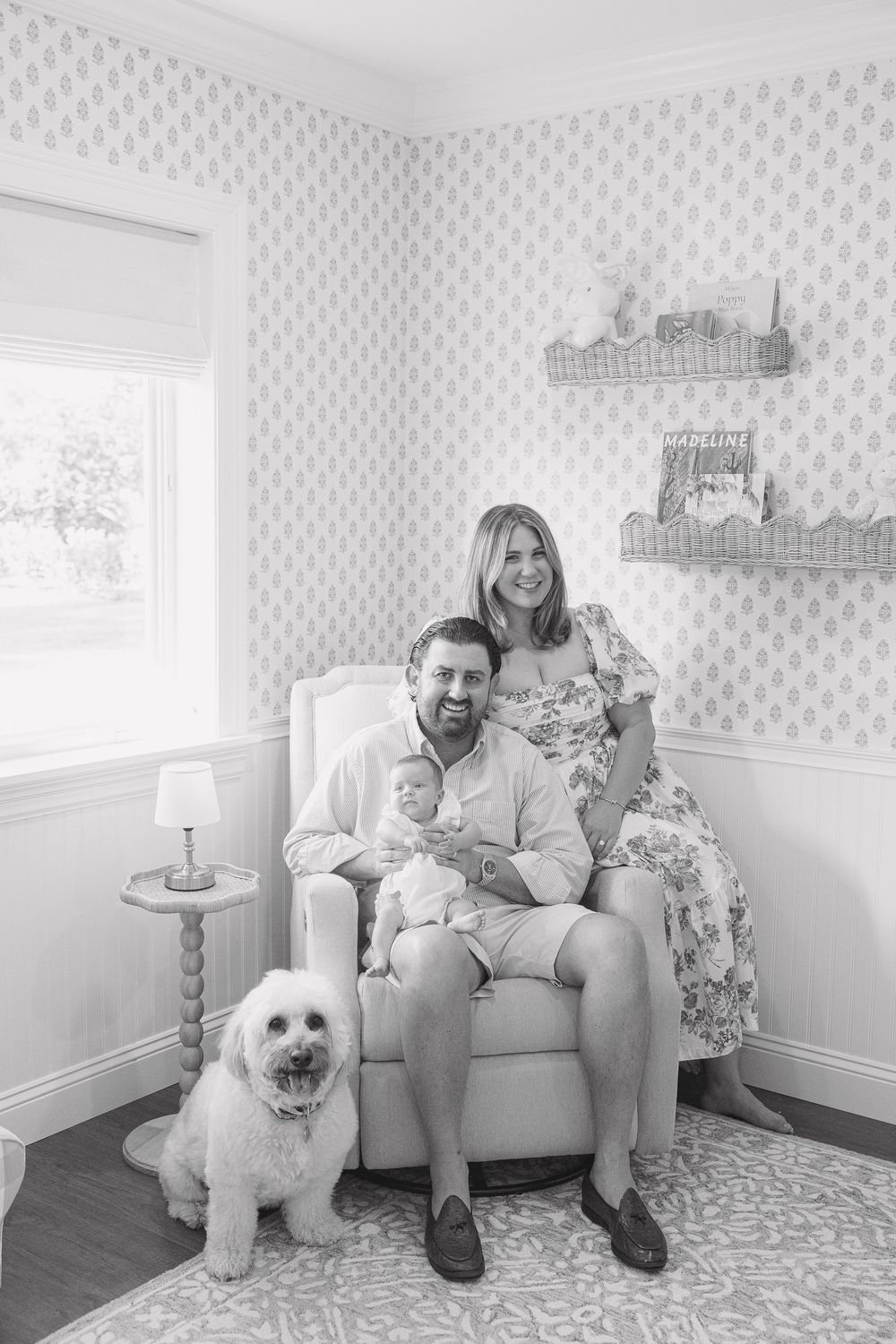 Black and white family portrait with parents, newborn baby, and fluffy white dog in a decorated nursery.
