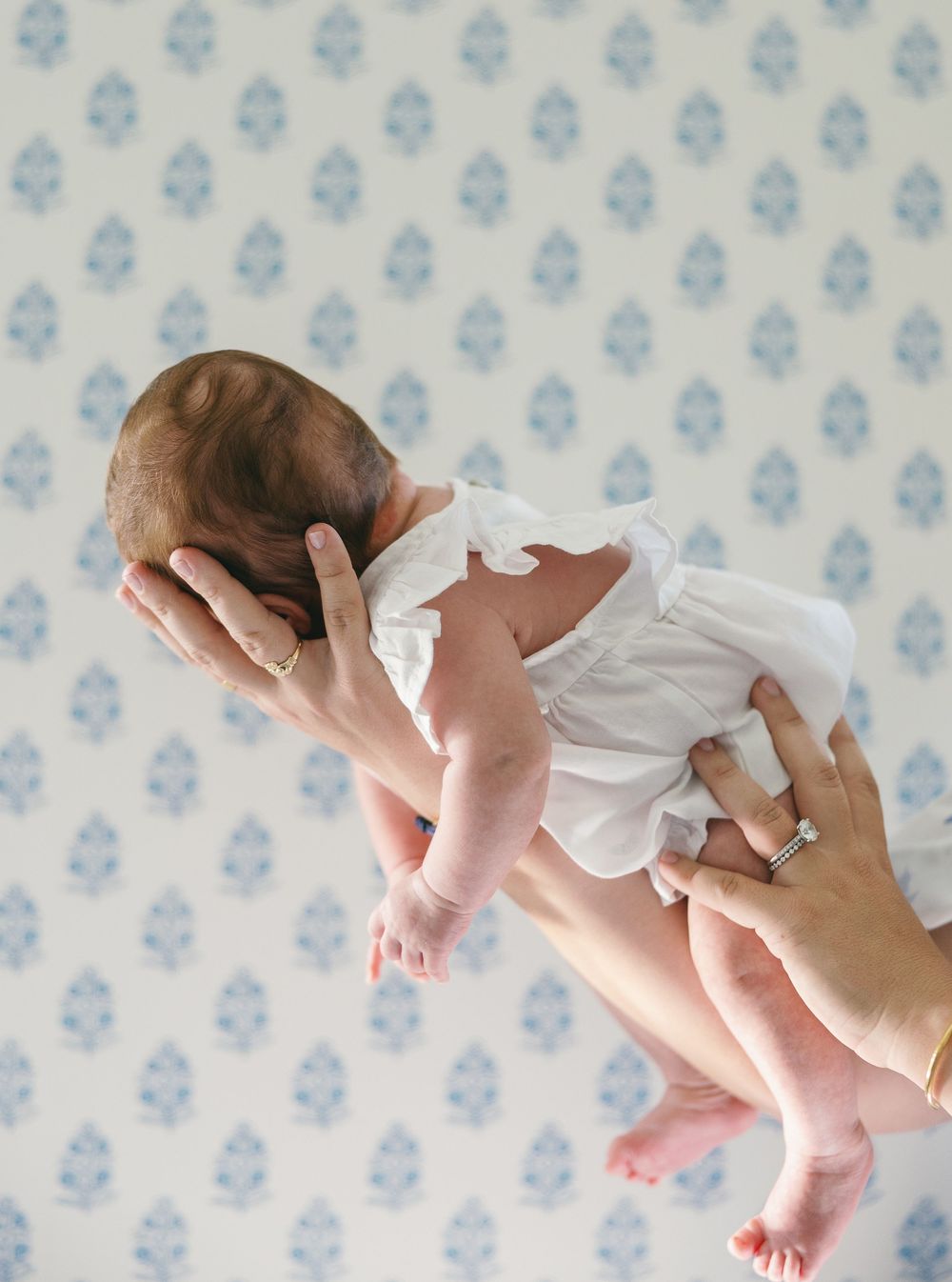 Adult hands gently holding a newborn baby in a white outfit against a blue floral wallpaper background.