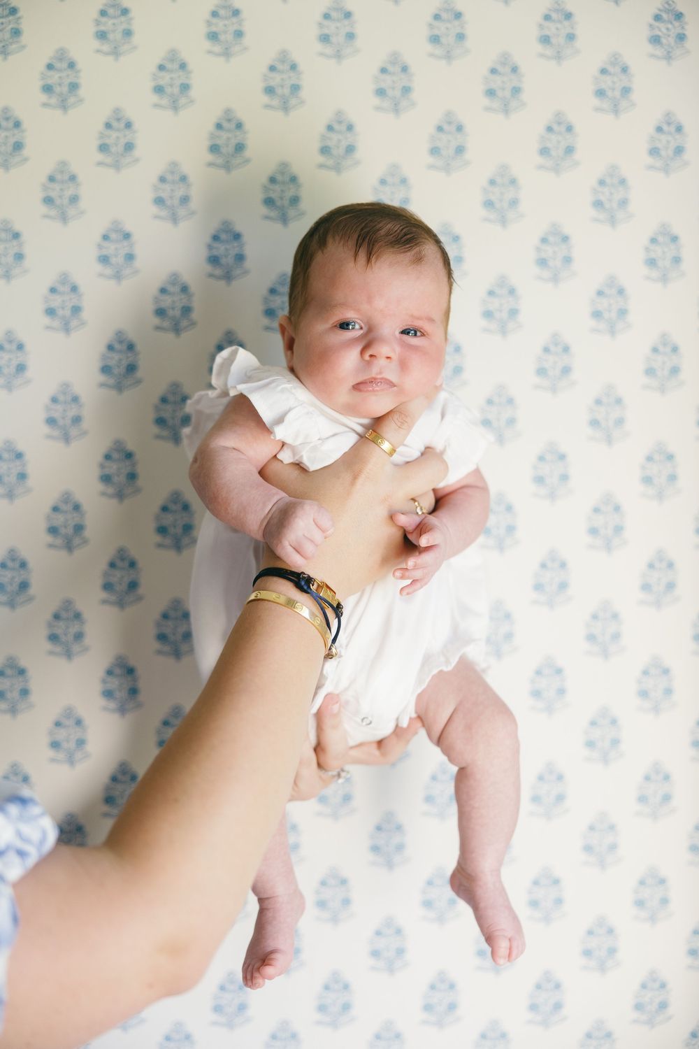 A newborn baby in a white outfit held up by an adult's hands against a blue floral wallpaper background.
