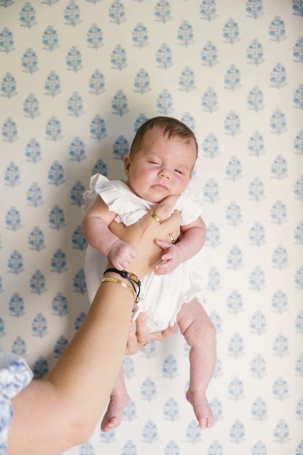 Newborn baby in white outfit held up against blue floral wallpaper during professional photo session.