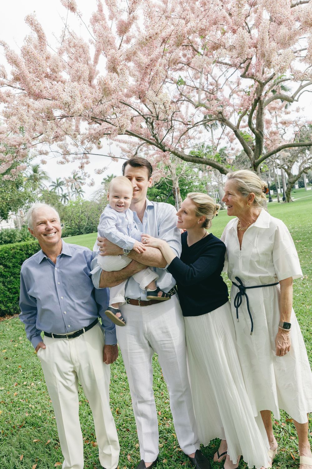 Multi-generational family posing outdoors under a blooming pink cherry blossom tree, including a baby.