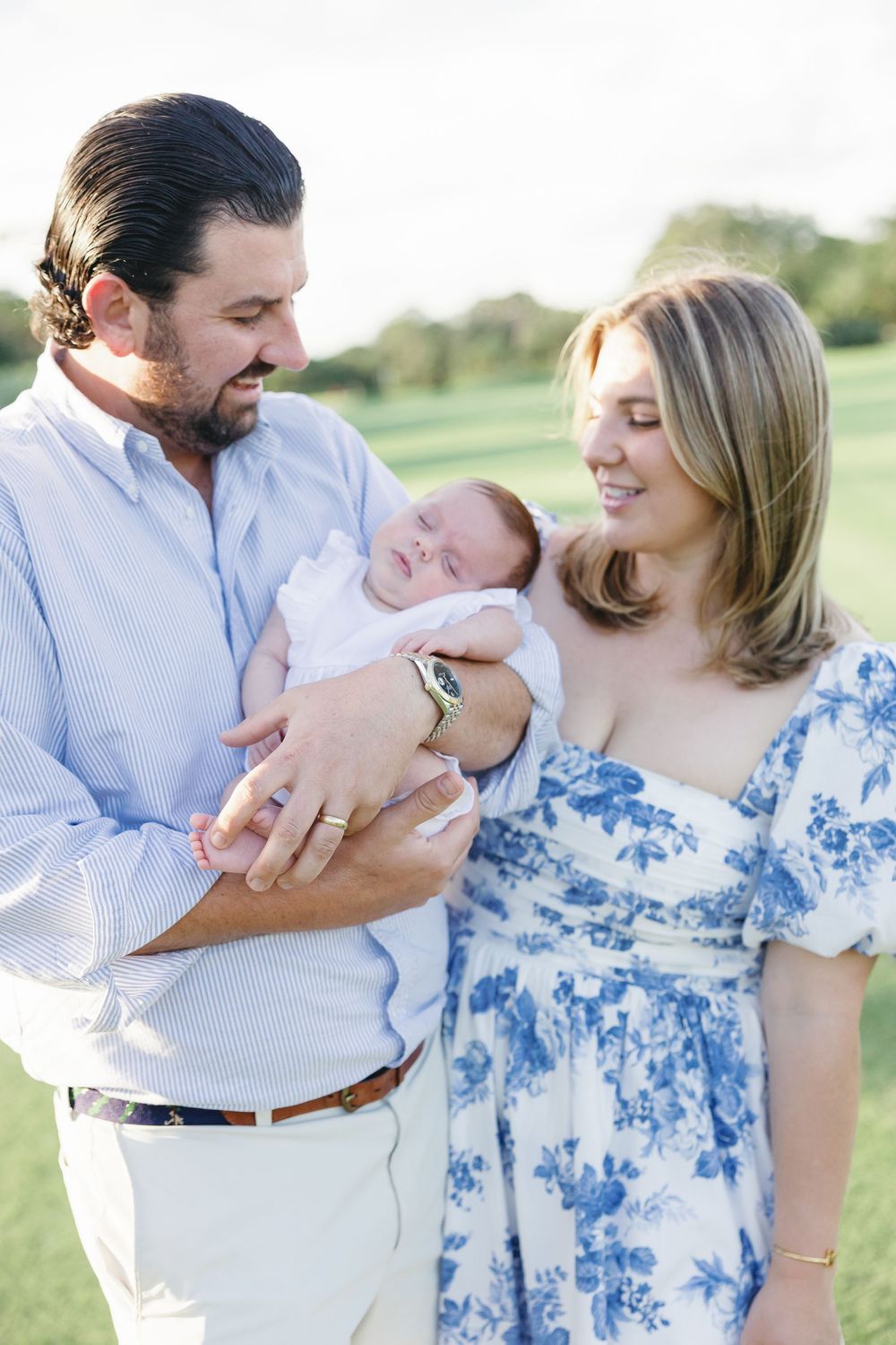 New parents tenderly gaze at their sleeping newborn baby held in dads arms outdoors.