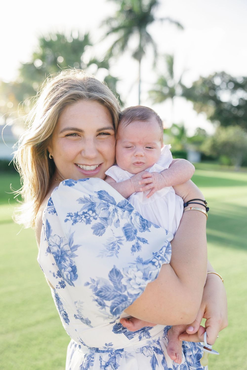 Smiling mother in blue floral dress holds sleeping newborn baby outdoors near palm trees.