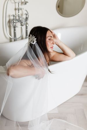 A bride in a white wedding veil relaxes in a luxurious freestanding bathtub during a bridal boudoir photoshoot.