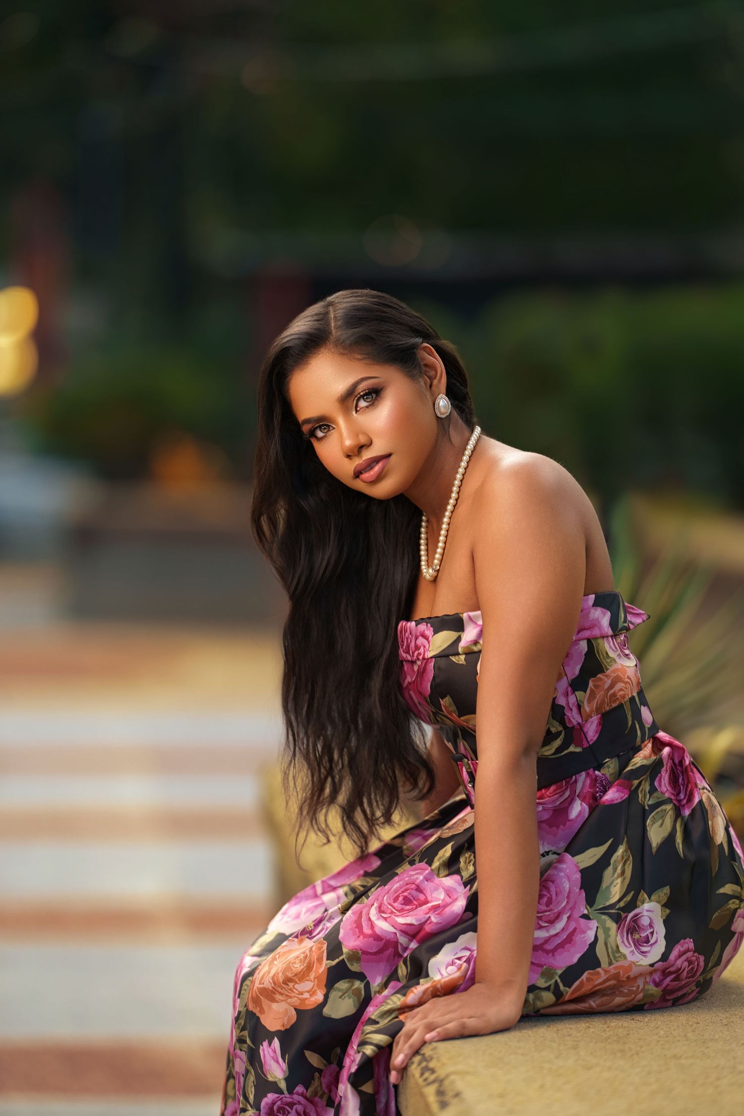 A woman in a floral print dress with long dark hair sits gracefully on stone steps at dusk.