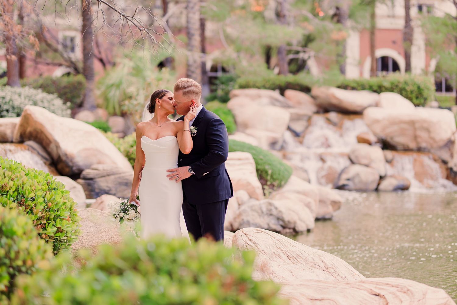 A couple shares a romantic embrace outdoors during their wedding, with the bride wearing a flowing veil in a natural setting.