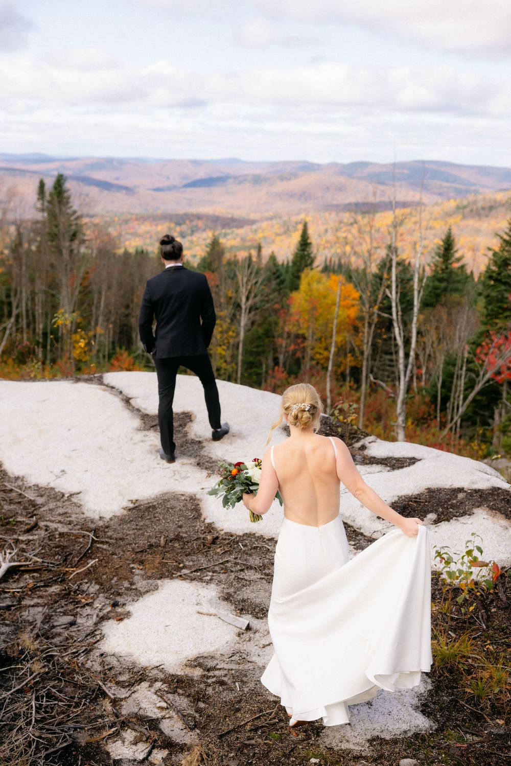 A couple stands on a snowy mountain peak overlooking a scenic autumn forest landscape with colorful fall foliage.
