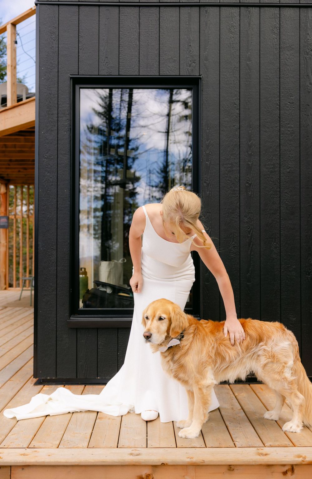 Golden retriever dog relaxes on a wooden deck next to a dark-colored exterior wall with window reflections.