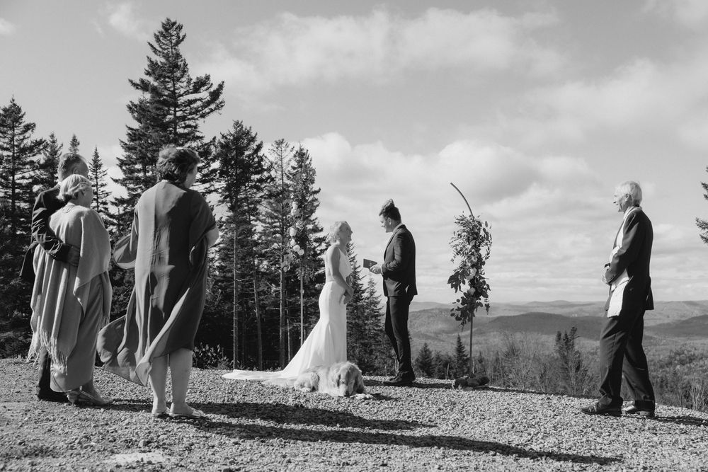A black and white wedding ceremony takes place on a scenic mountaintop with evergreen trees in the background.