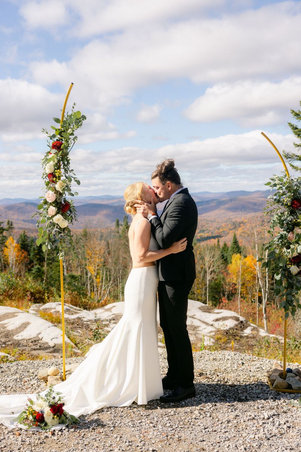A romantic outdoor mountain wedding ceremony with a couple embracing against a backdrop of autumn foliage and distant peaks.