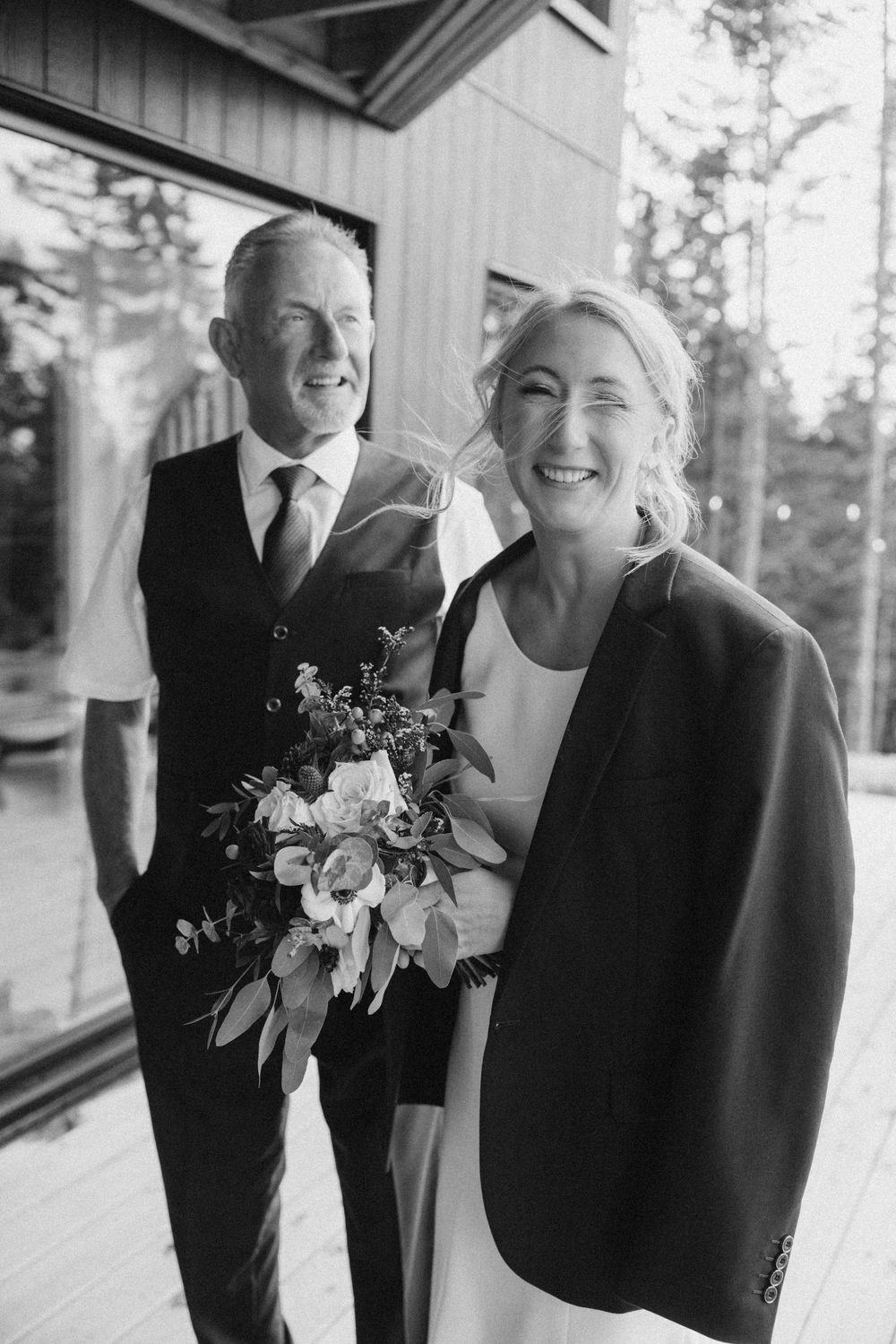 A joyful wedding couple shares candid laughter while posing together on a covered porch.