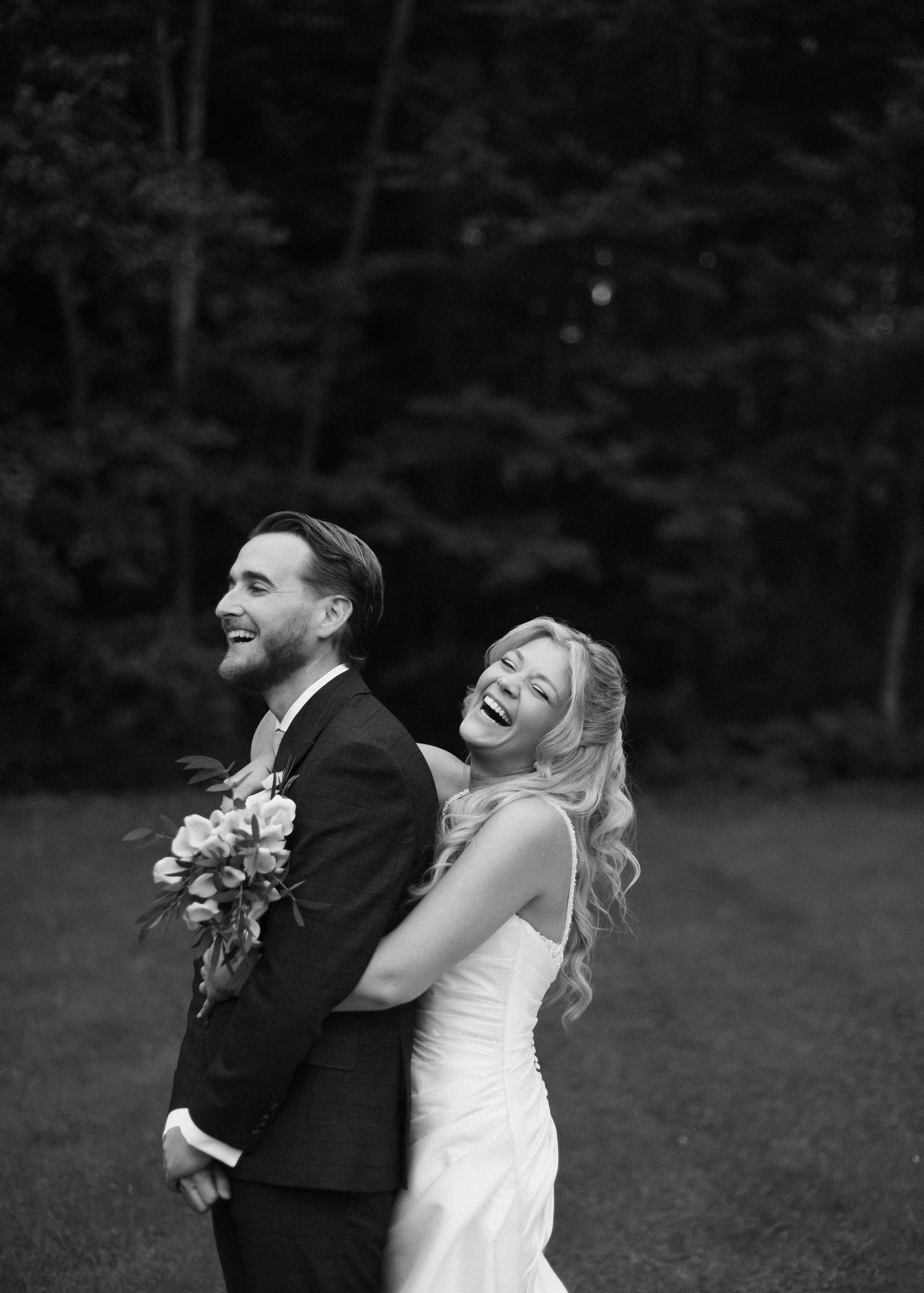 Black and white photo of a joyful bride and groom laughing together outdoors during their wedding celebration.