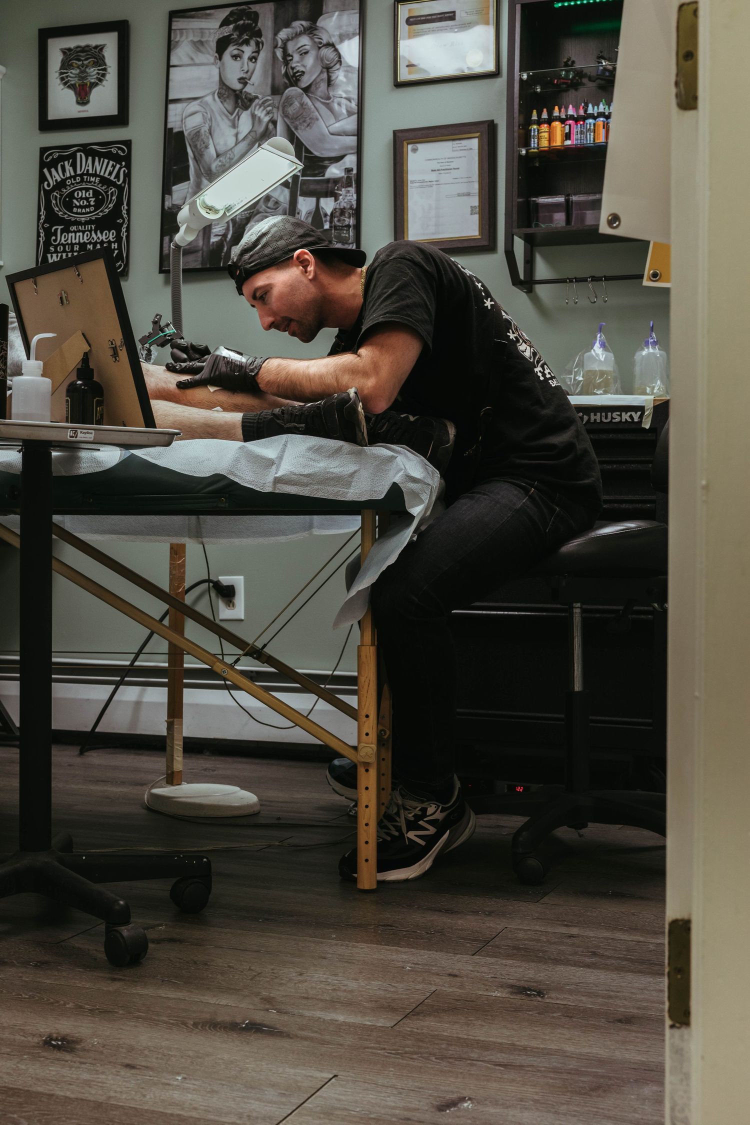 Tattoo artist working at station in vintage-styled shop with framed artwork on walls.