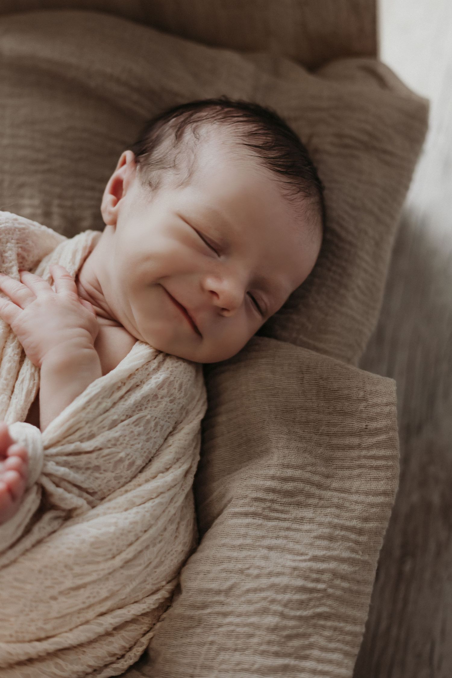 Peaceful sleeping newborn wrapped in soft beige muslin blanket resting on neutral toned bedding.