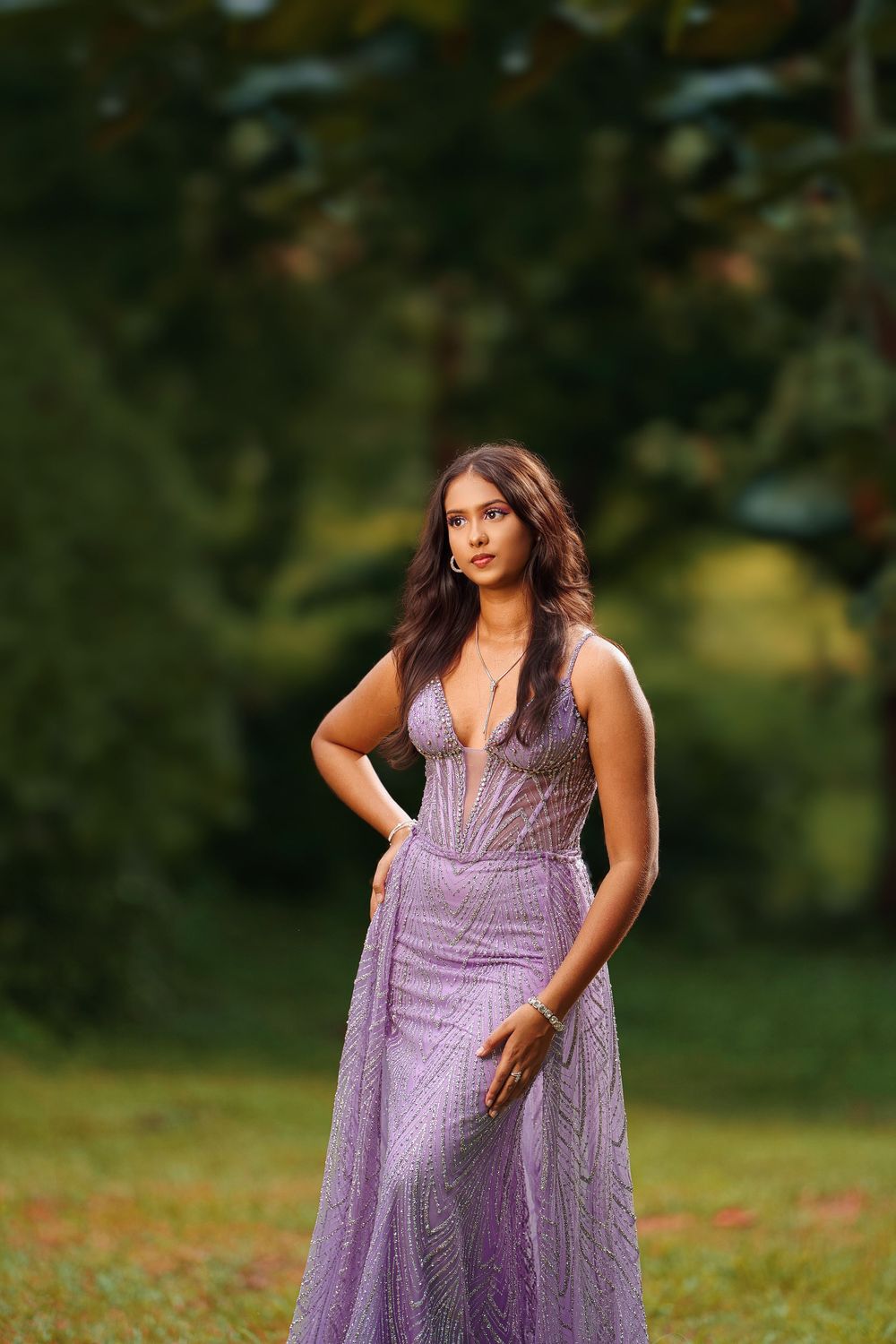 A woman poses in a flowing lavender evening gown outdoors in a natural garden setting during golden hour.