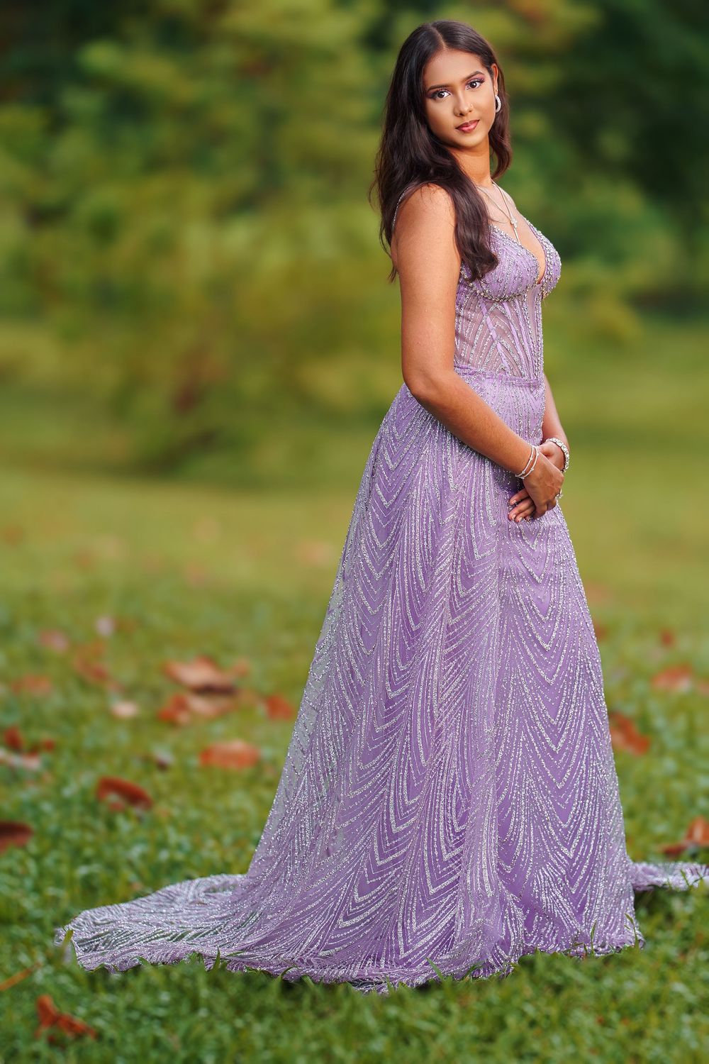 A person in a flowing lavender lace gown poses gracefully in a garden setting with autumn leaves scattered on the grass.