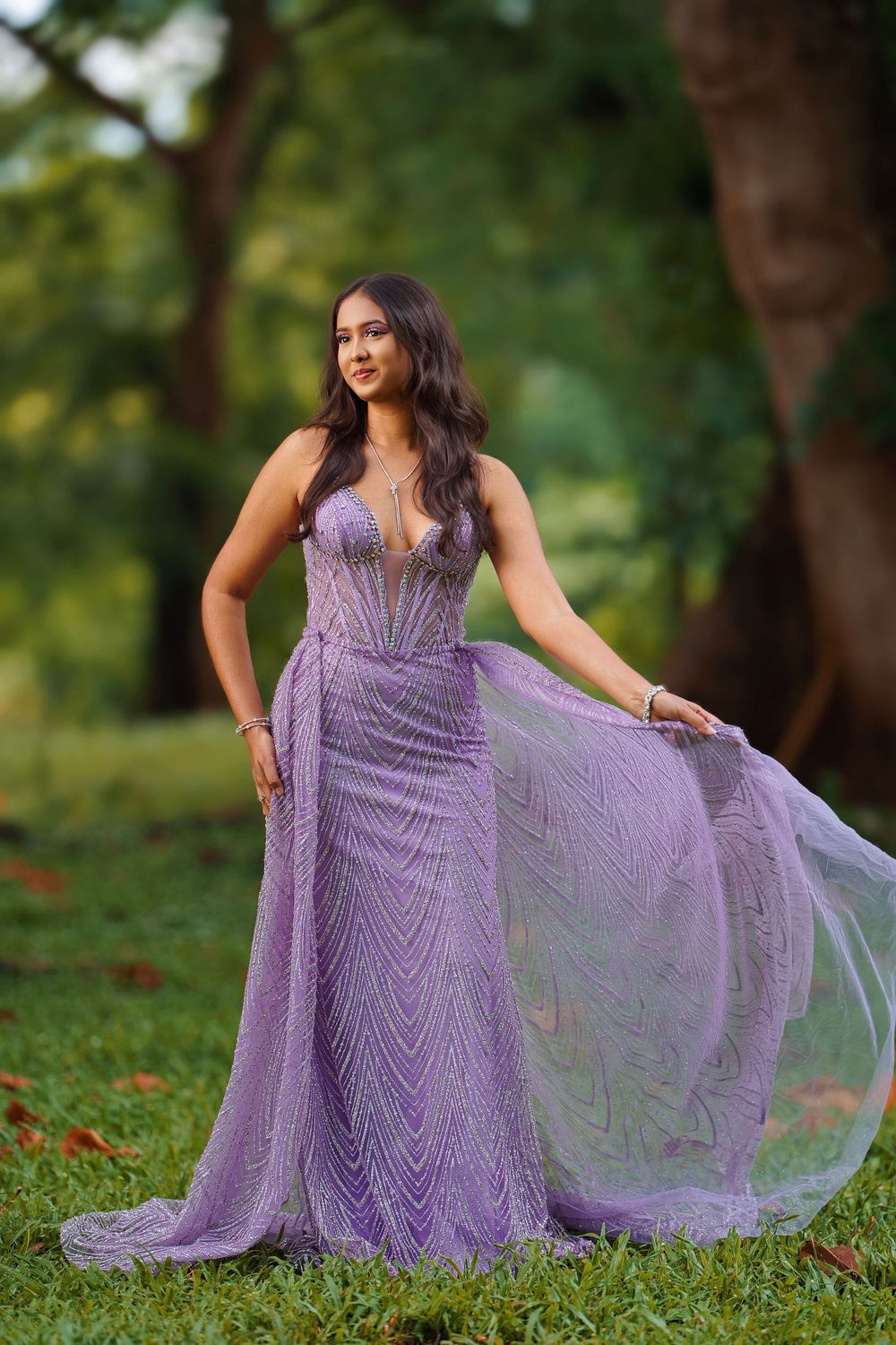 A person in a flowing lavender gown with beaded details poses gracefully on grass in an outdoor garden setting.